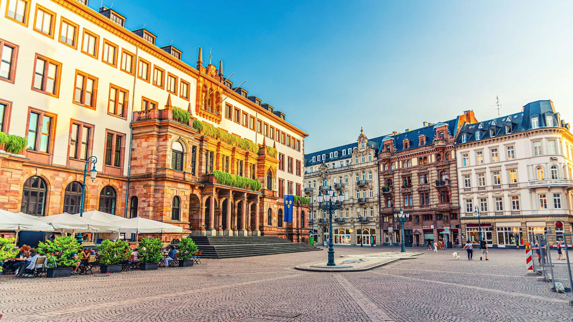 "A vibrant, sunlit view of the historic city center of Wiesbaden, Germany, featuring the grand Old Town Hall with a terraced cafe and colorful buildings in the background.  "