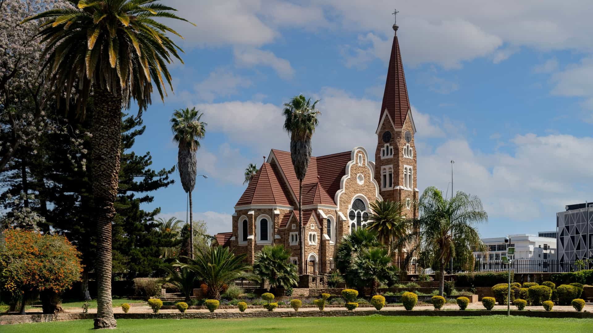 The Christ Church is a neo-Romanesque church with a red steeple, set in a green garden with towering palm trees.