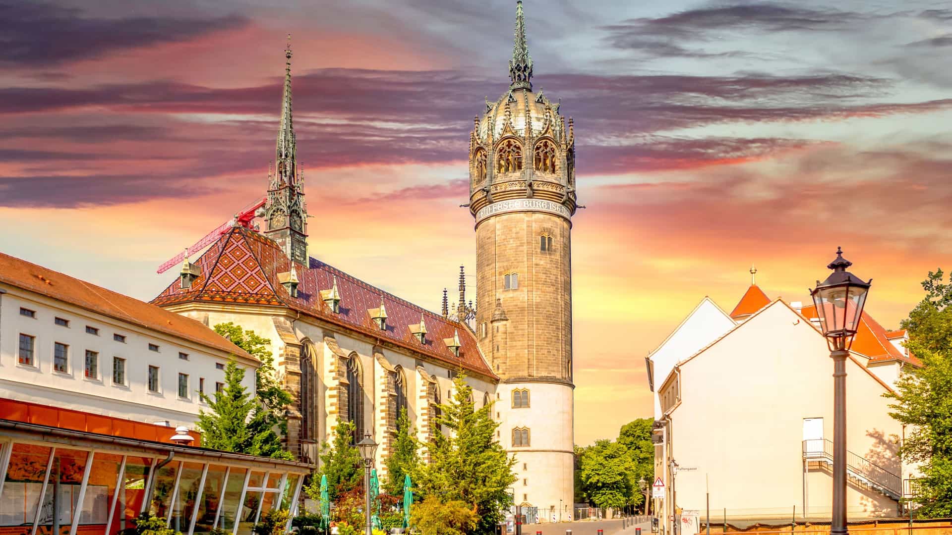 "A breathtaking sunset over the historic Wittenberg All Saints' Church in Germany, with its ornate tower and spires against a sky filled with dramatic pink and orange clouds.  "