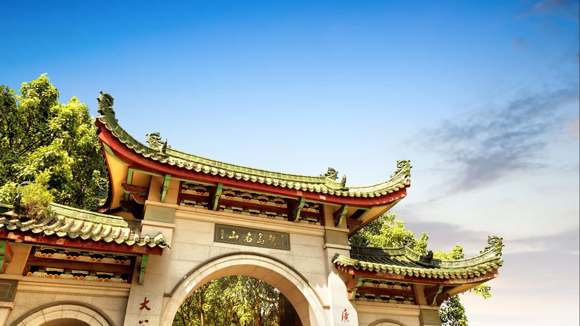 A low-angle view of a traditional Chinese archway, a "paifang," with a green, curved roof and ornate carvings under a bright blue sky, a landmark in Xiamen.