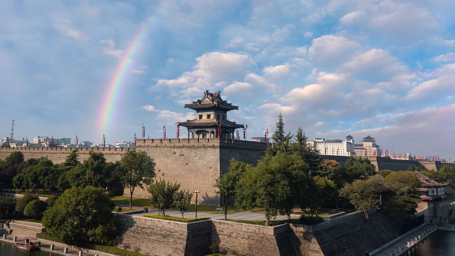 A vibrant rainbow arcs over the historic City Wall in Xi'an, China, a large defensive stone structure with a traditional Chinese watchtower and surrounding moat.