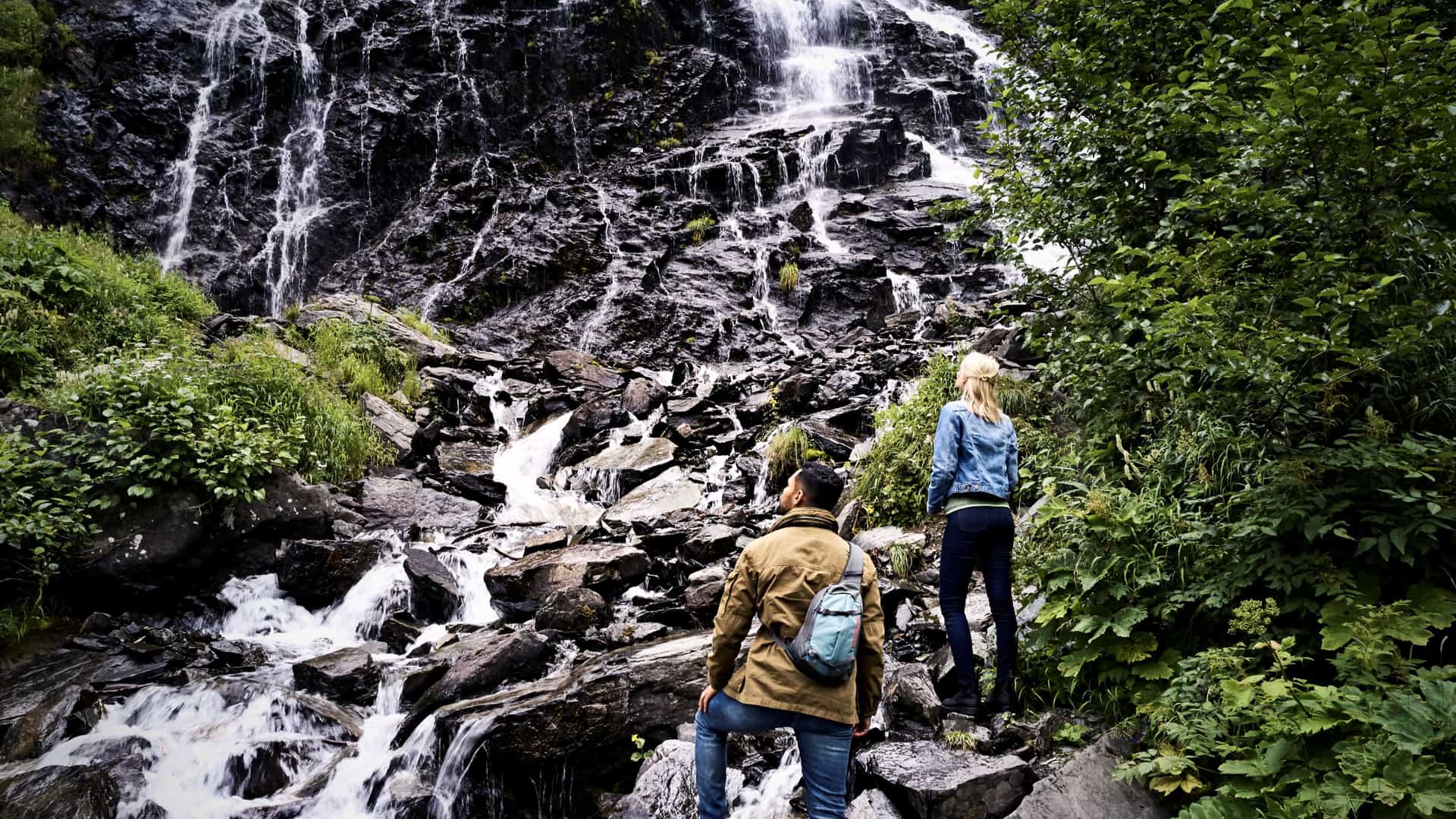 Two travelers explore a rocky trail at the base of a cascading waterfall in a lush Alaskan forest.
