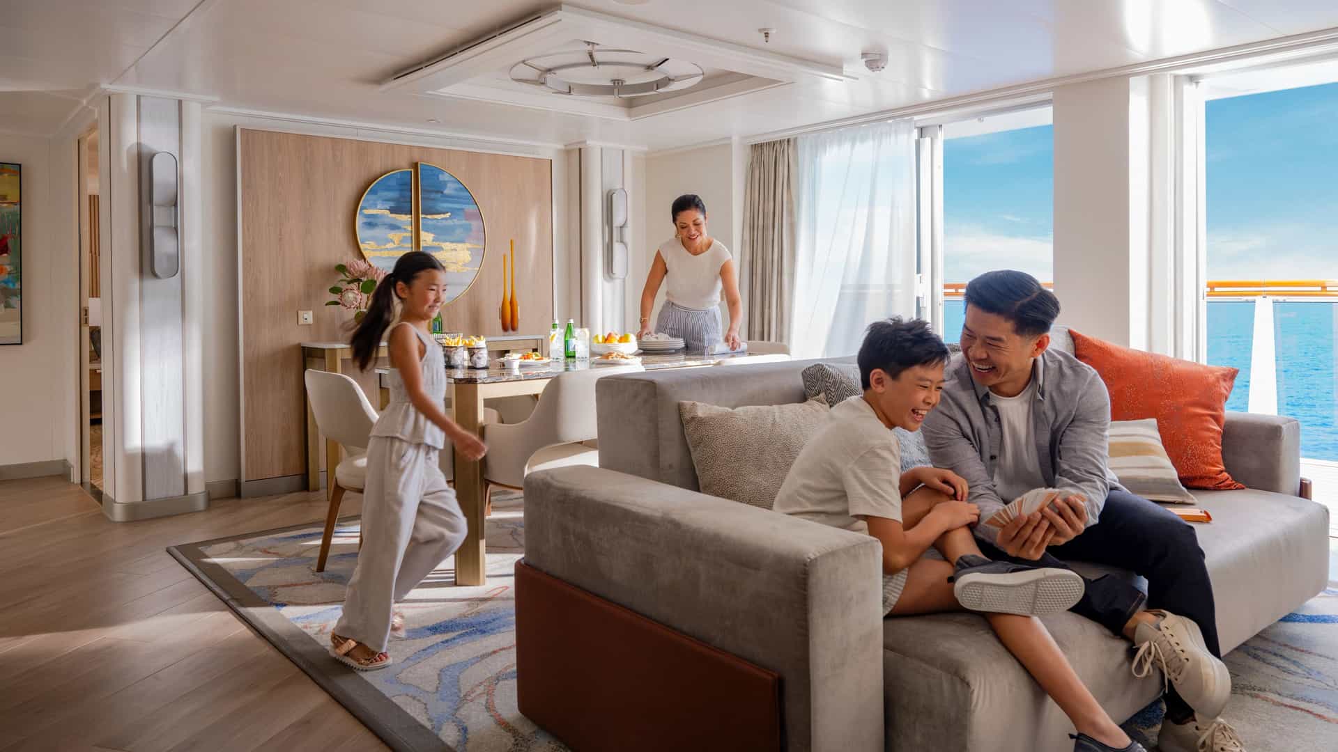An Asian family relaxing inside a spacious Sky Suite onboard a Sun Princess cruise ship, with a father and son playing cards on the couch and a mother and daughter near the dining area by a balcony window overlooking the ocean.