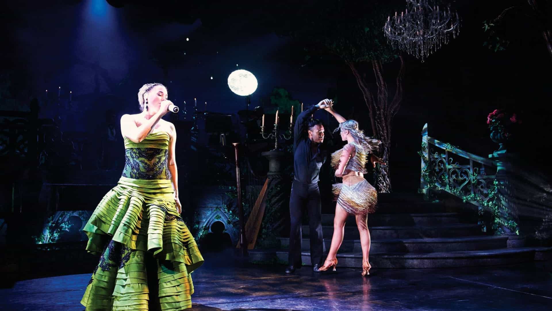 A dynamic theatrical performance on a cruise ship stage. A female singer in a green dress sings into a microphone, while a male and female dancer perform a routine in the background. The stage is set with a staircase and a large chandelier, creating a dramatic, mystical atmosphere. The image suggests a new form of entertainment and repositioning of the cruise experience.