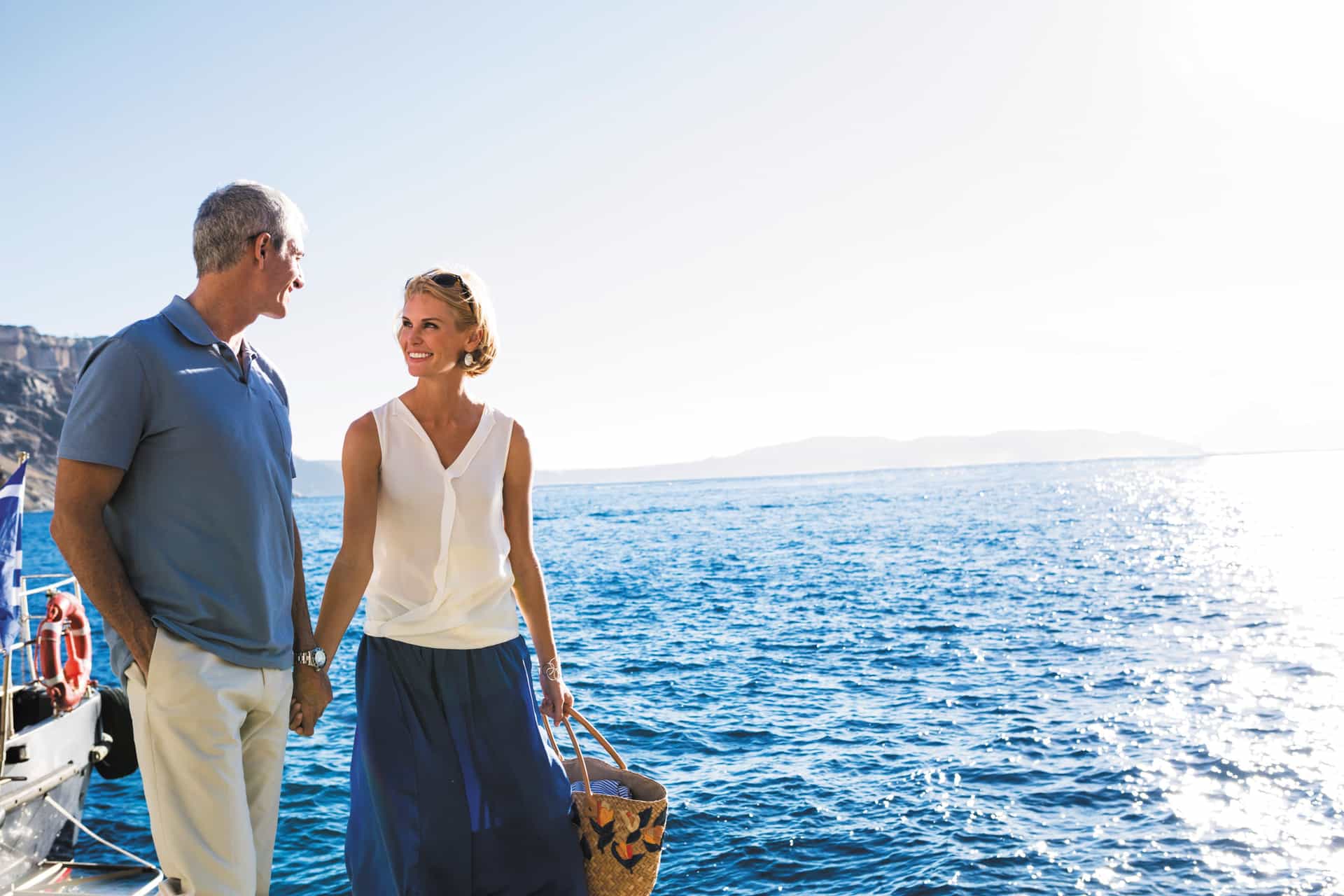 A smiling mature couple holds hands on a boat deck, enjoying a scenic cruise on the bright blue sea with the coast of Santorini visible in the distance.