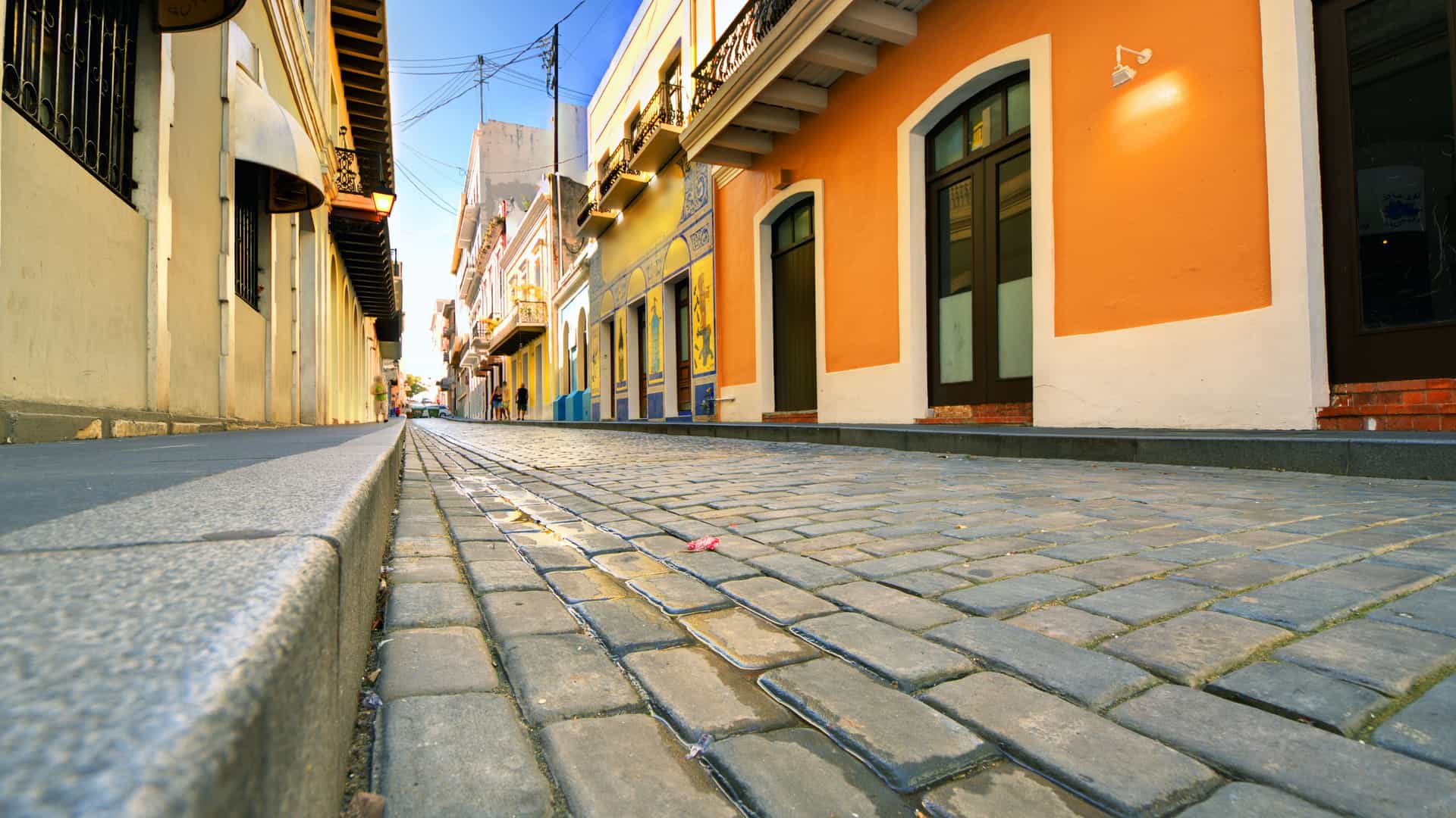 A low-angle, close-up view of a historic cobblestone street in Old San Juan, Puerto Rico. The blue-gray cobblestones, known as adoquines, create a textured pathway, with the low sun casting long shadows. The street is lined with brightly colored colonial buildings, painted in shades of orange, yellow, and white, with balconies and arched doorways. The scene captures the charming, historic atmosphere of this vibrant city in the Southern Caribbean.