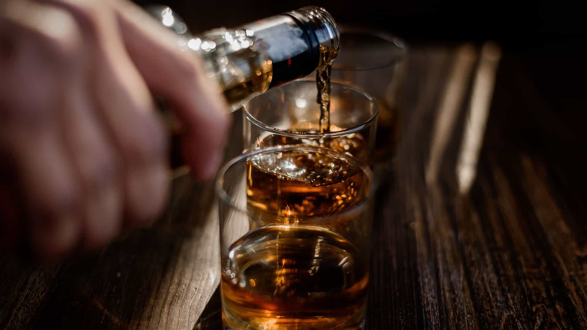 A hand pours dark rum from a bottle into a row of three small glasses on a rustic wooden table. The amber liquid splashes into the glasses, which are arranged diagonally in the foreground. The warm color of the rum and the soft, natural light creates a cozy atmosphere. The image suggests a rum tasting or a social occasion, highlighting the rum production that is a key part of the culture and history of the Southern Caribbean island of Barbados, home to the Mount Gay Rum distillery.