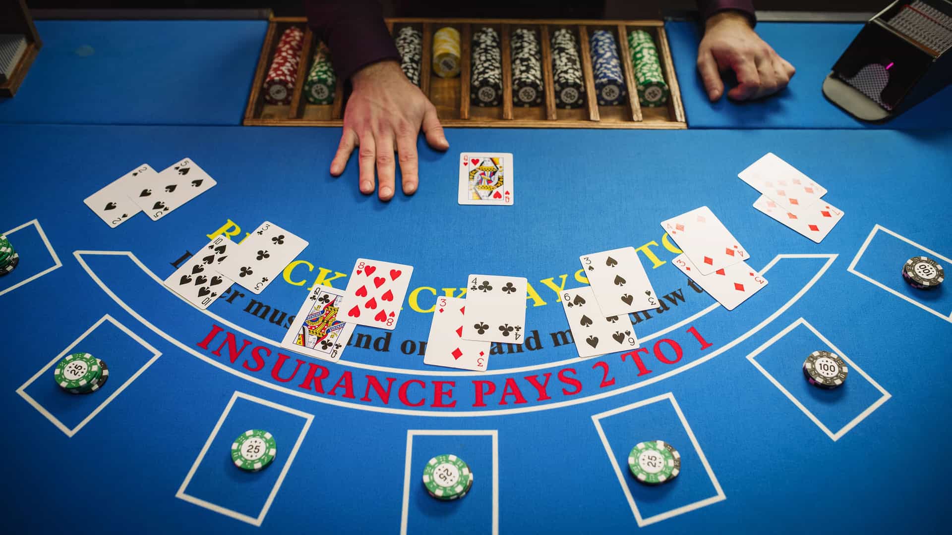 A close-up, elevated view of a blackjack table in a casino in Aruba. The table is covered in blue felt with the word 'blackjack' and other rules printed on it. There are several hands of cards laid out, with some showing winning hands and others losing. A pair of hands, likely the dealer's, is visible at the top of the frame, along with a wooden rack of colorful casino chips. The image captures the vibrant and exciting casino nightlife that is a popular attraction on the Southern Caribbean island of Aruba.