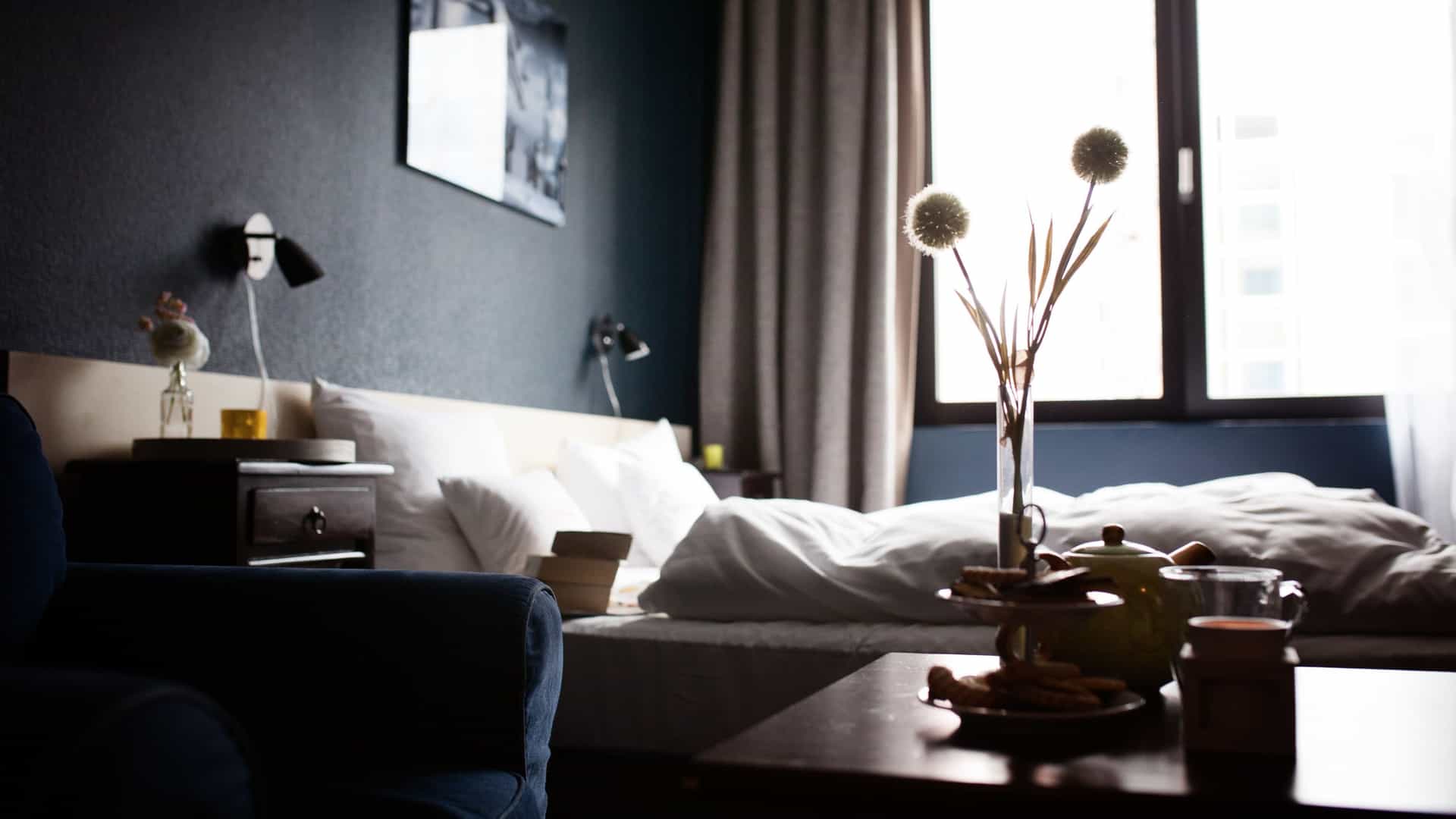 A dimly lit, cozy hotel room featuring a bed with white linens, dark blue walls, and a foreground table set with tea and snacks in front of a bright window.
