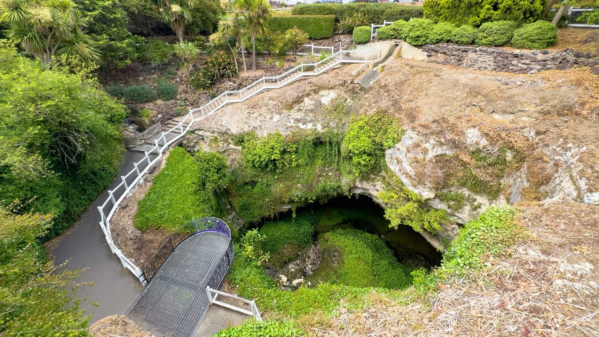 A landscaped Mt. Gambier sinkhole with lush greenery, white‑railed pathways, and a metal bridge spanning the cave opening beneath bright garden light.