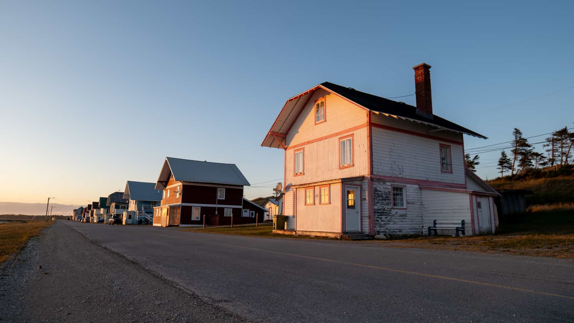 A sunlit Port Menier street with a weathered white‑and‑red wooden house, quiet roadside homes, parked vehicles, and warm coastal evening light over Anticosti Island.