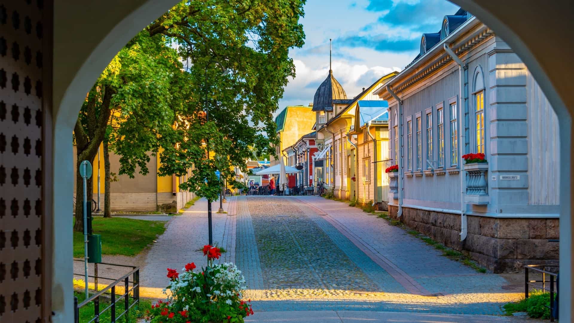 A cobblestone street in Old Rauma framed by an archway, with colorful wooden houses, flower boxes, and warm sunlight leading toward outdoor café tables.