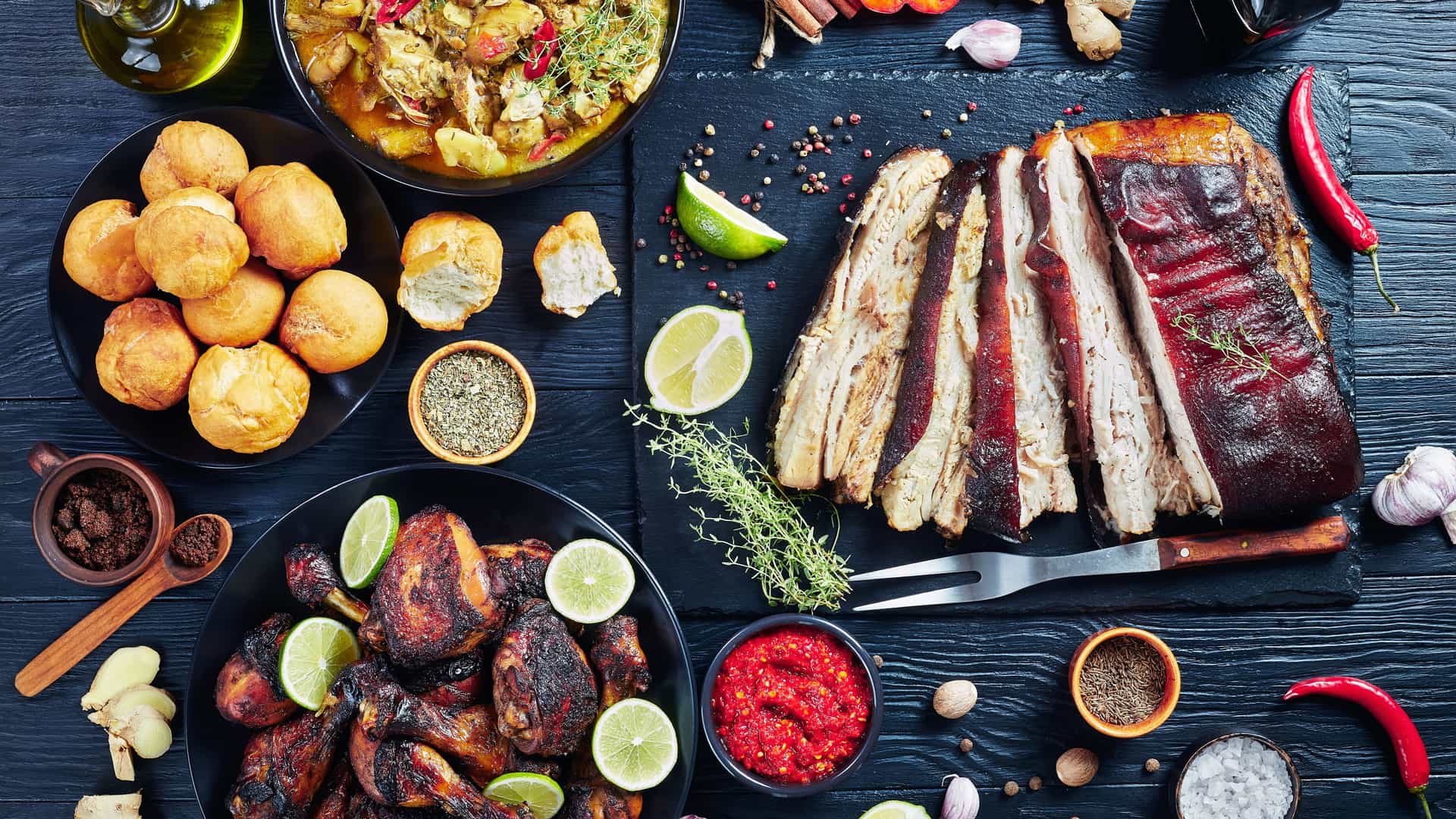 An overhead view of a feast of Western Caribbean cuisine. The table is laden with several dishes, including a plate of savory jerk chicken drumsticks garnished with lime wedges and a bowl of chicken curry. There is also a plate of golden fried dumplings, a cut of roasted pork belly, and various spices like thyme and allspice. Red peppers, fresh ginger, and garlic cloves are scattered on the dark wooden surface. The image captures the rich and diverse flavors of the Caribbean region.