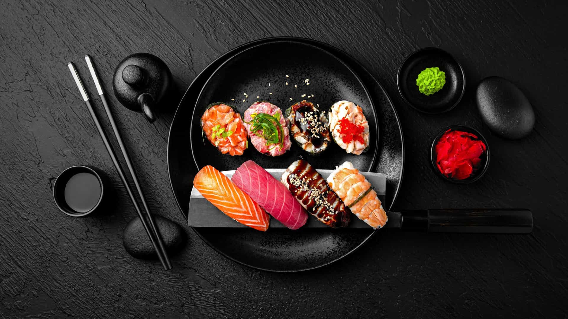 A beautiful, high-angle flat lay of a variety of sushi nigiri and rolls artfully arranged on a dark, textured surface. The plate holds several pieces of sushi, including nigiri with salmon and tuna, and rolls topped with eel and fish roe. Accompanying the sushi are small bowls containing green wasabi and pink pickled ginger, a small ceramic pot for soy sauce, and a pair of elegant black chopsticks. The image captures a classic example of exquisite Japanese cuisine.