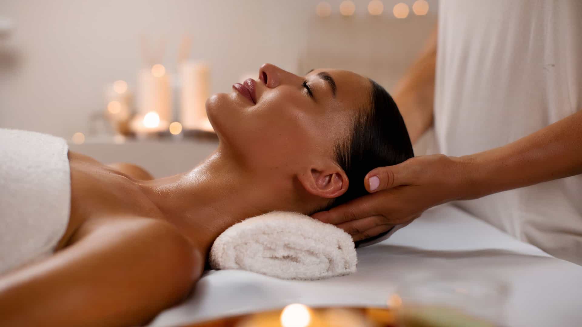 A serene scene of a woman relaxing during a head massage in a spa. She is lying on a massage table with her eyes closed, her hair pulled back, and her shoulders draped in a white towel. A professional massage therapist is gently cradling her head with both hands. The background is softly lit with several candles, creating a warm and tranquil atmosphere. The image captures the relaxing and luxurious experience often found on cruise ships and in spas around the world.