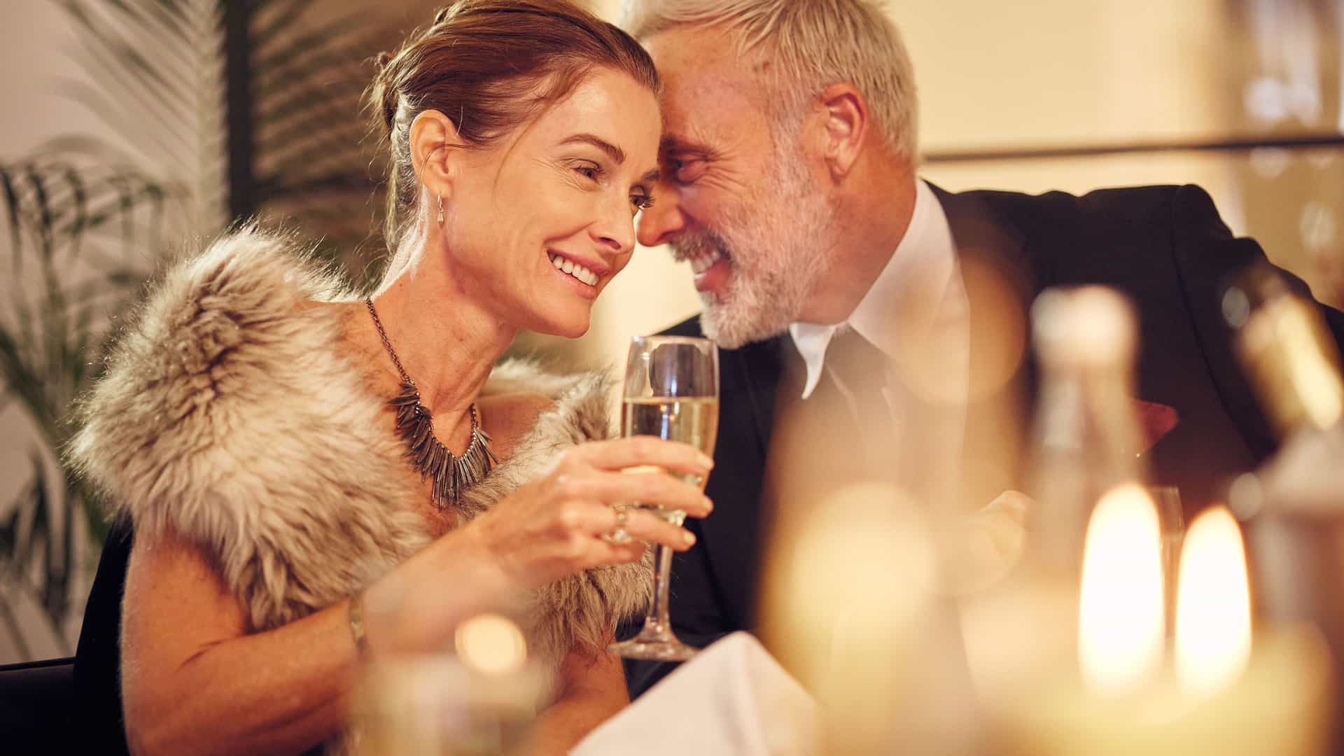 A stylish, mature couple enjoying a romantic evening at a fine dining restaurant. The woman, with her hair in an updo and wearing a fur shawl, holds a glass of champagne and smiles warmly at the man. The man, dressed in a formal suit, is leaning in and laughing with her, their heads close together. The table is set with wine glasses, napkins, and lit candles, creating a soft, intimate ambiance. The image captures a special celebration, a common theme in luxury travel and cruising.