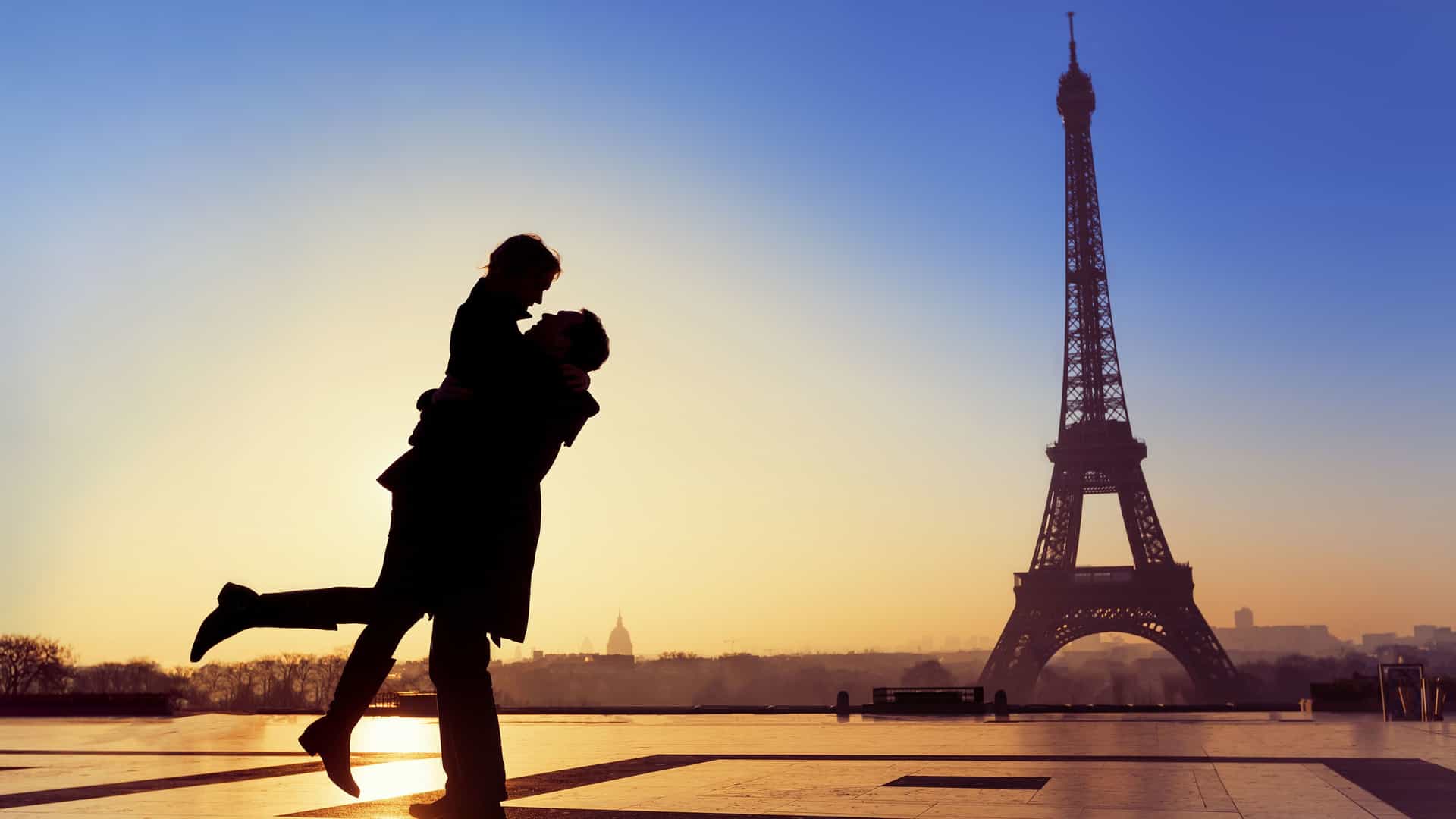 A silhouette of a romantic couple embracing in front of the Eiffel Tower in Paris, France. The man is lifting the woman off the ground, and they are looking into each other's eyes. Their figures are dark against the bright, golden glow of the setting sun. The Eiffel Tower stands tall and dark against the gradient of the evening sky, and the city skyline is faintly visible in the background. The image captures the romantic atmosphere of a world-famous landmark.