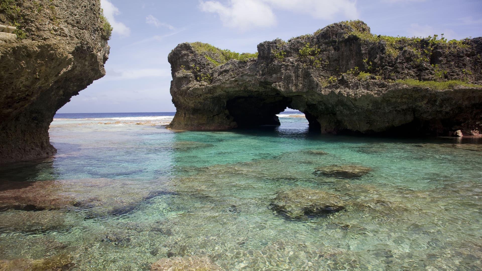 A stunning natural rock pool with an archway carved into the rock, leading to the open ocean, on the island of Alofi. The water is clear and shallow, with a rocky bottom visible beneath the surface.