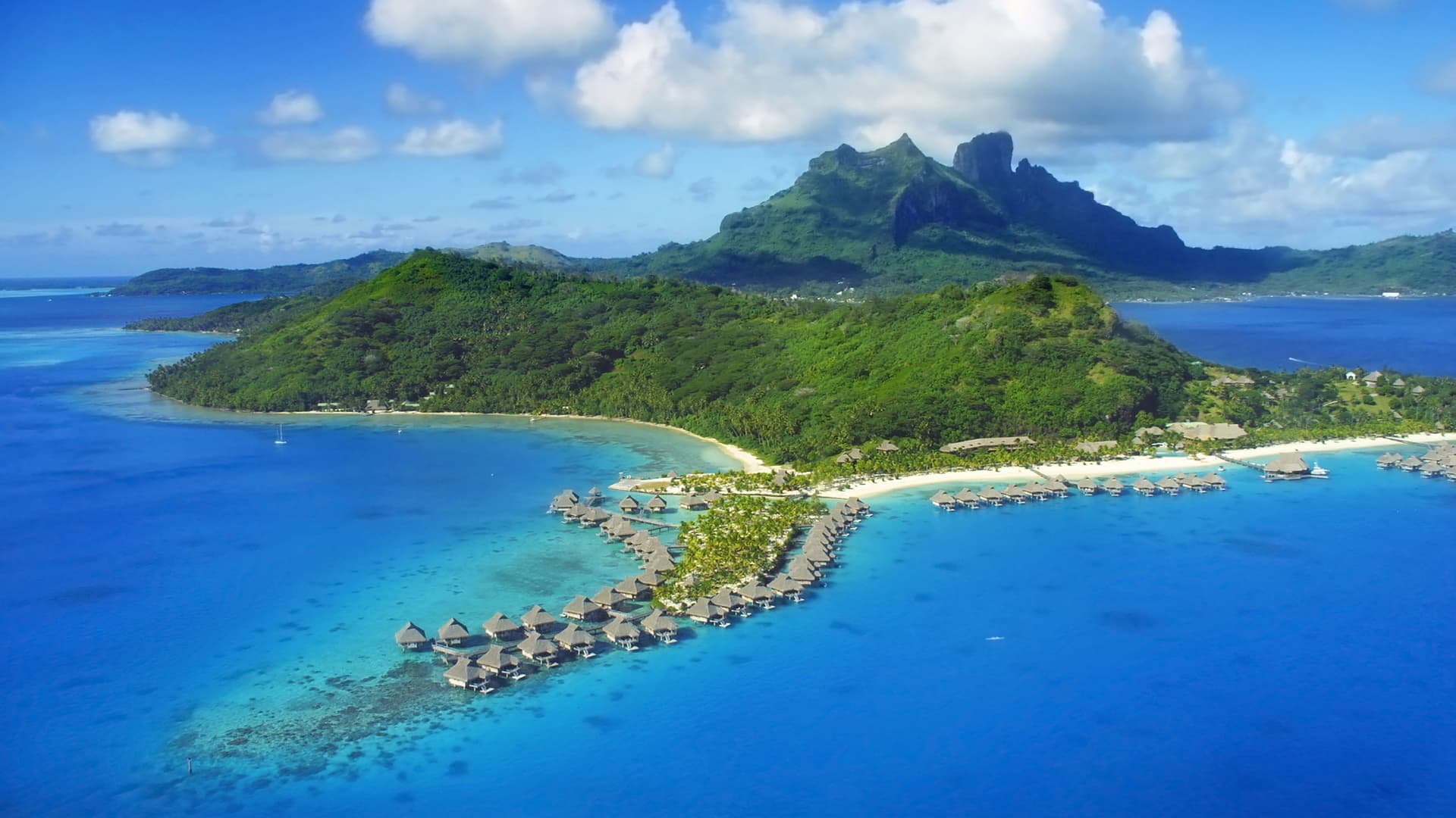 An aerial view of Bora Bora, a South Pacific island with lush green, volcanic peaks, and turquoise blue waters, surrounded by luxury overwater bungalows on a coral reef.