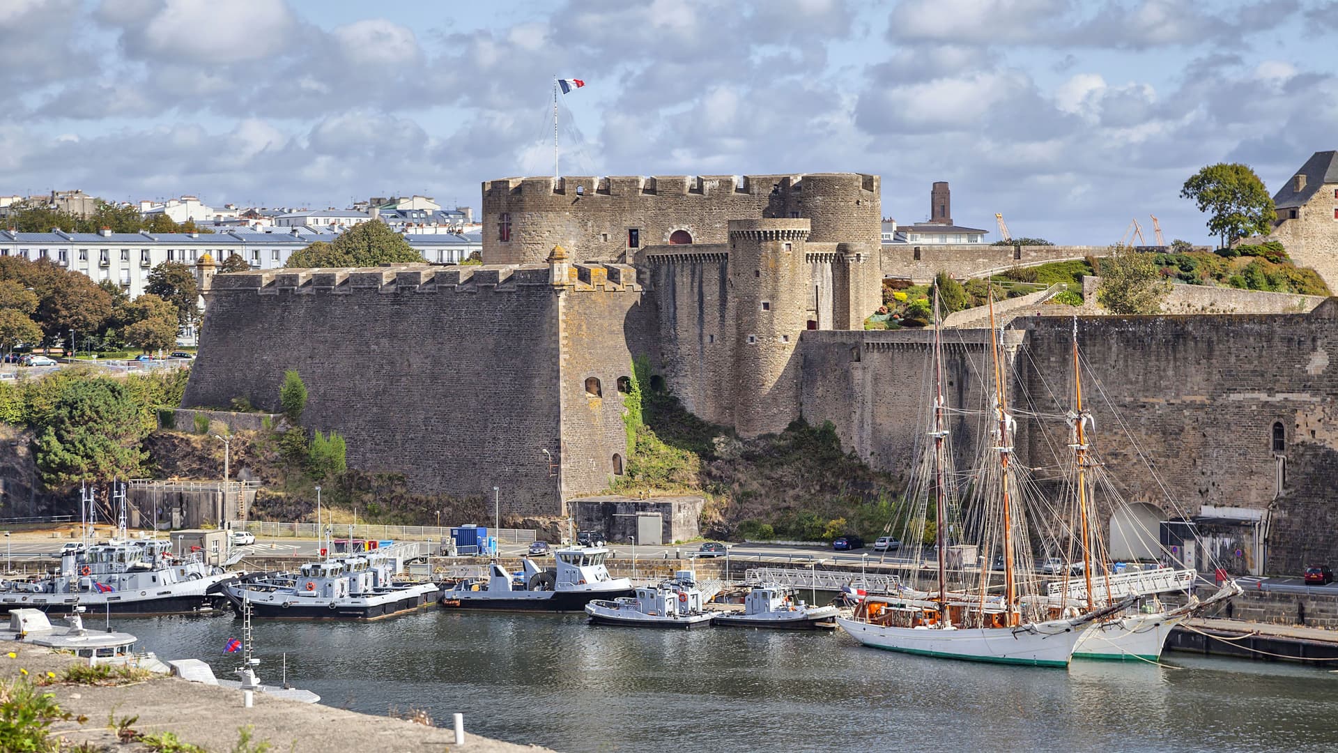 A beautiful port scene in Brest, France, with modern boats and a large sailboat docked in the foreground, and the historic medieval fortress, Château de Brest, towering over the harbor under a blue sky.
