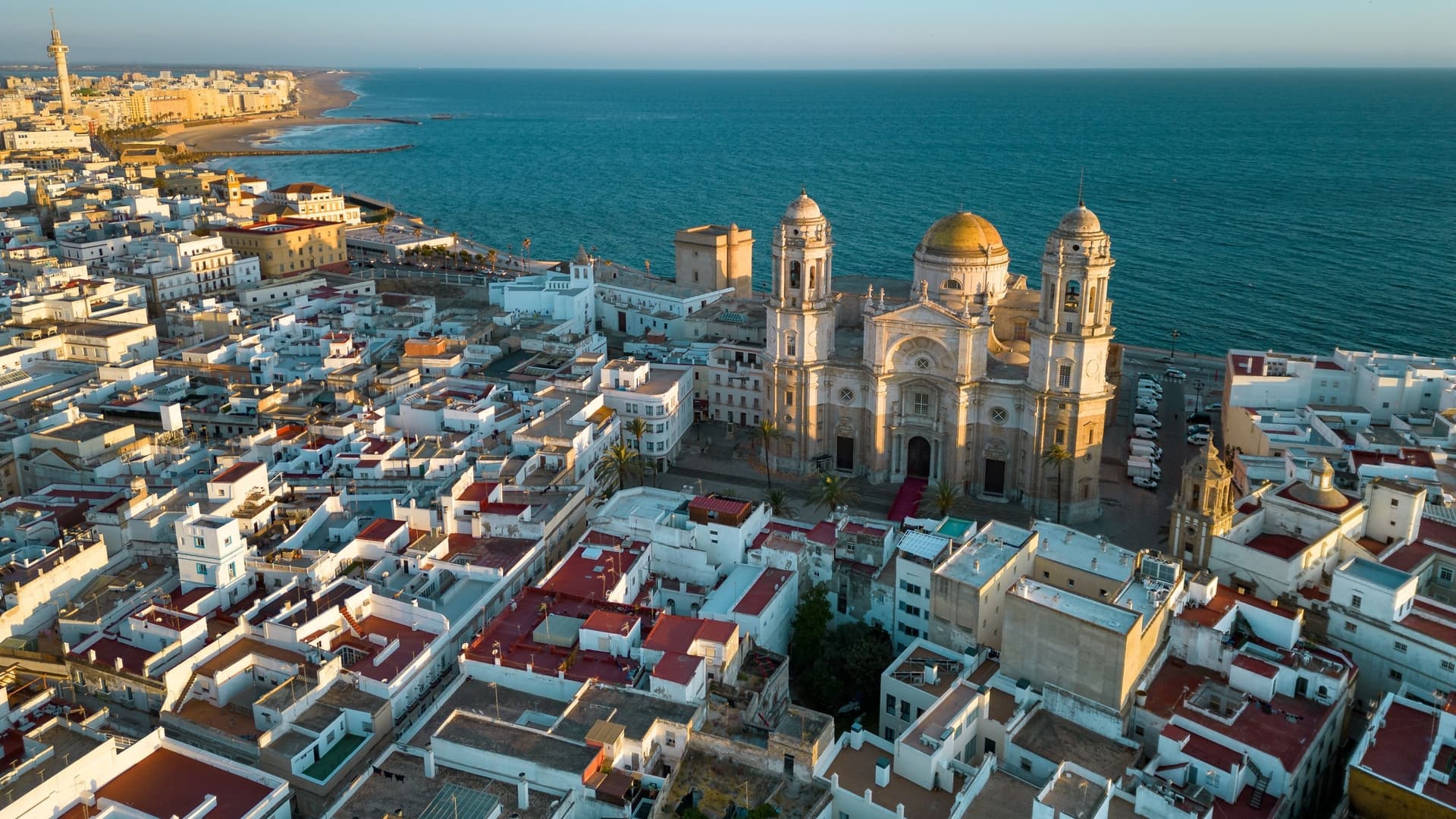 An aerial, drone-perspective photograph of the city center of Cadiz, Spain, captured at sunset. The baroque and neoclassical Cathedral of Cadiz, with its distinctive golden dome and bell towers, is prominently centered among the densely packed, white and warm-toned buildings of the historic old town. Warm, golden sunlight reflects off the rooftops, highlighting the medieval layout of the city. The vast blue expanse of the Atlantic Ocean is visible in the background.