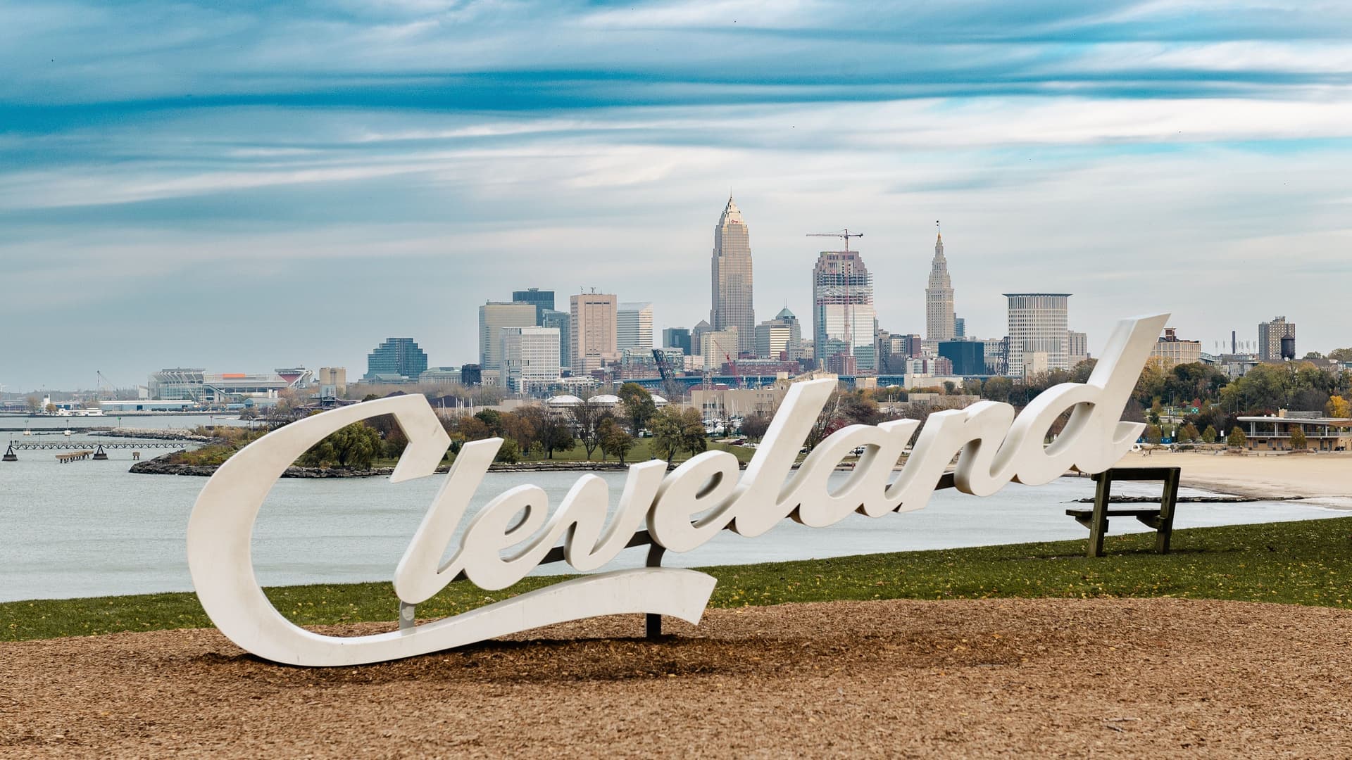 A panoramic shot of the Cleveland skyline, with its downtown skyscrapers and Lake Erie in the background, featuring the large white "Cleveland" sign in the foreground.