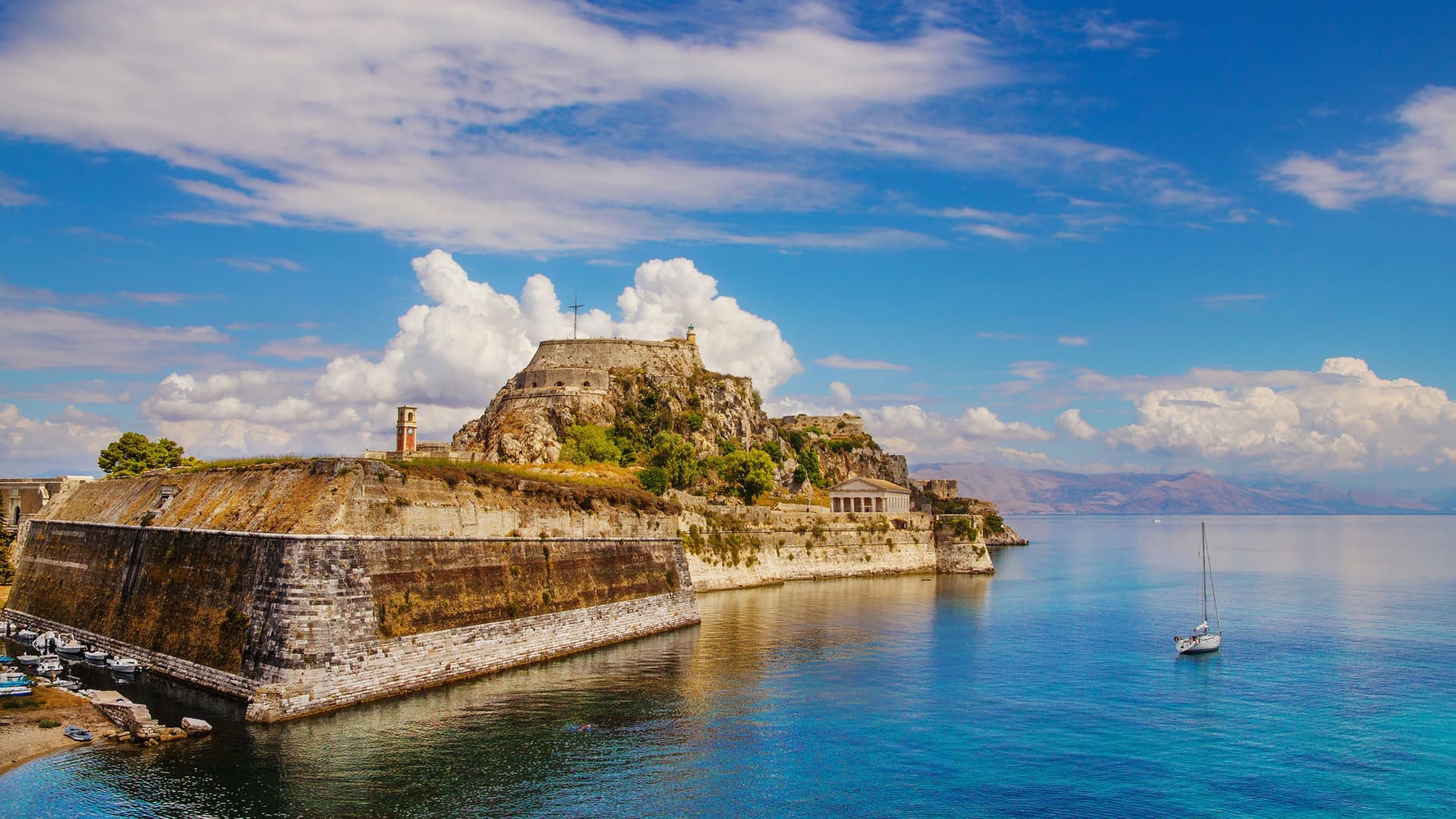 A picturesque view of the historic Old Fortress of Corfu, Greece, with its stone walls rising from the turquoise Ionian Sea and a sailboat in the foreground.