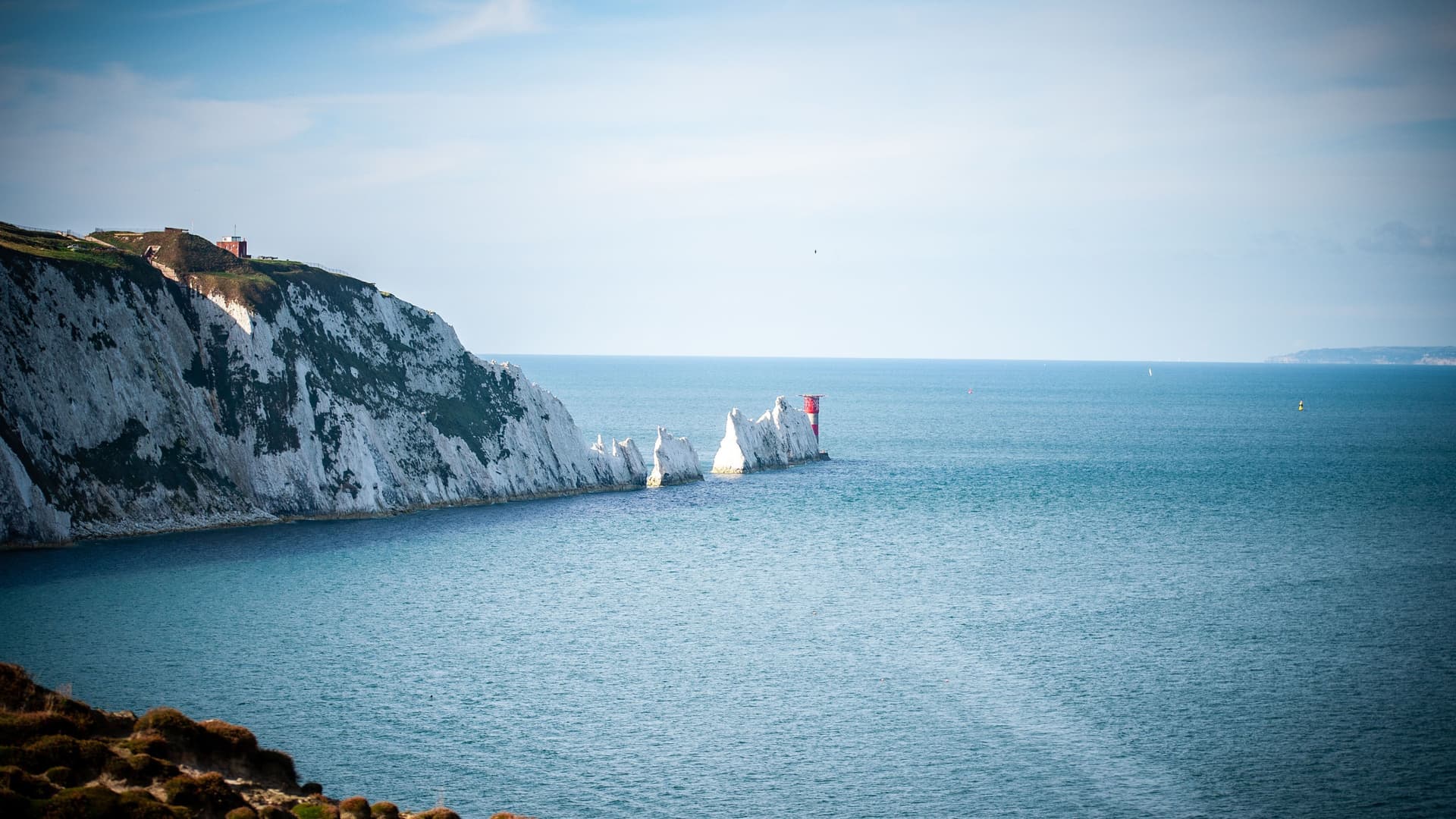 A scenic view of The Needles, iconic chalk stacks with a red and white striped lighthouse, extending from the white cliffs of the Isle of Wight into the blue sea.