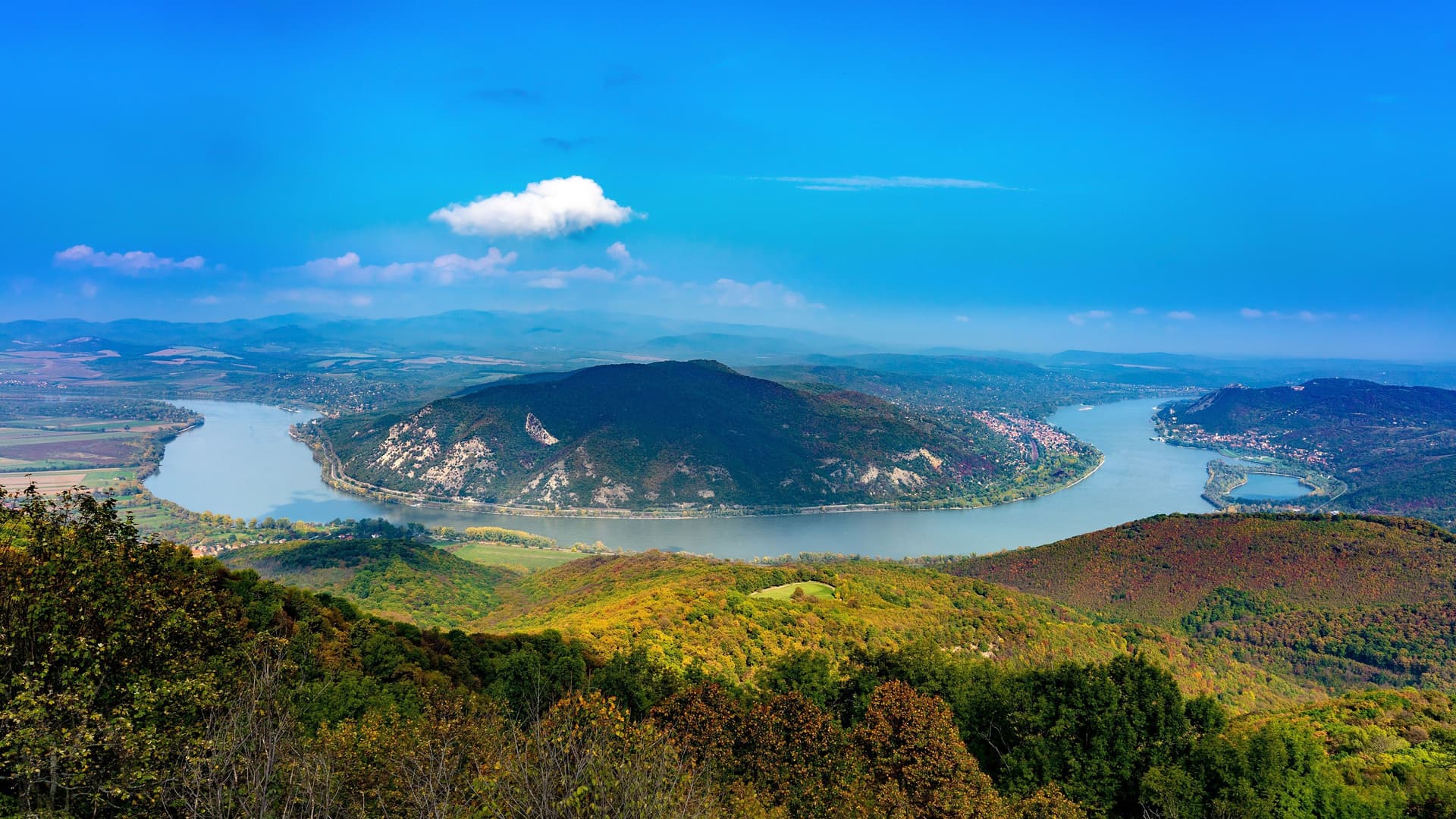 A wide-angle panoramic view of the dramatic horseshoe-shaped Danube Bend, a famous landmark in Hungary, surrounded by autumn-colored forests and a charming town.