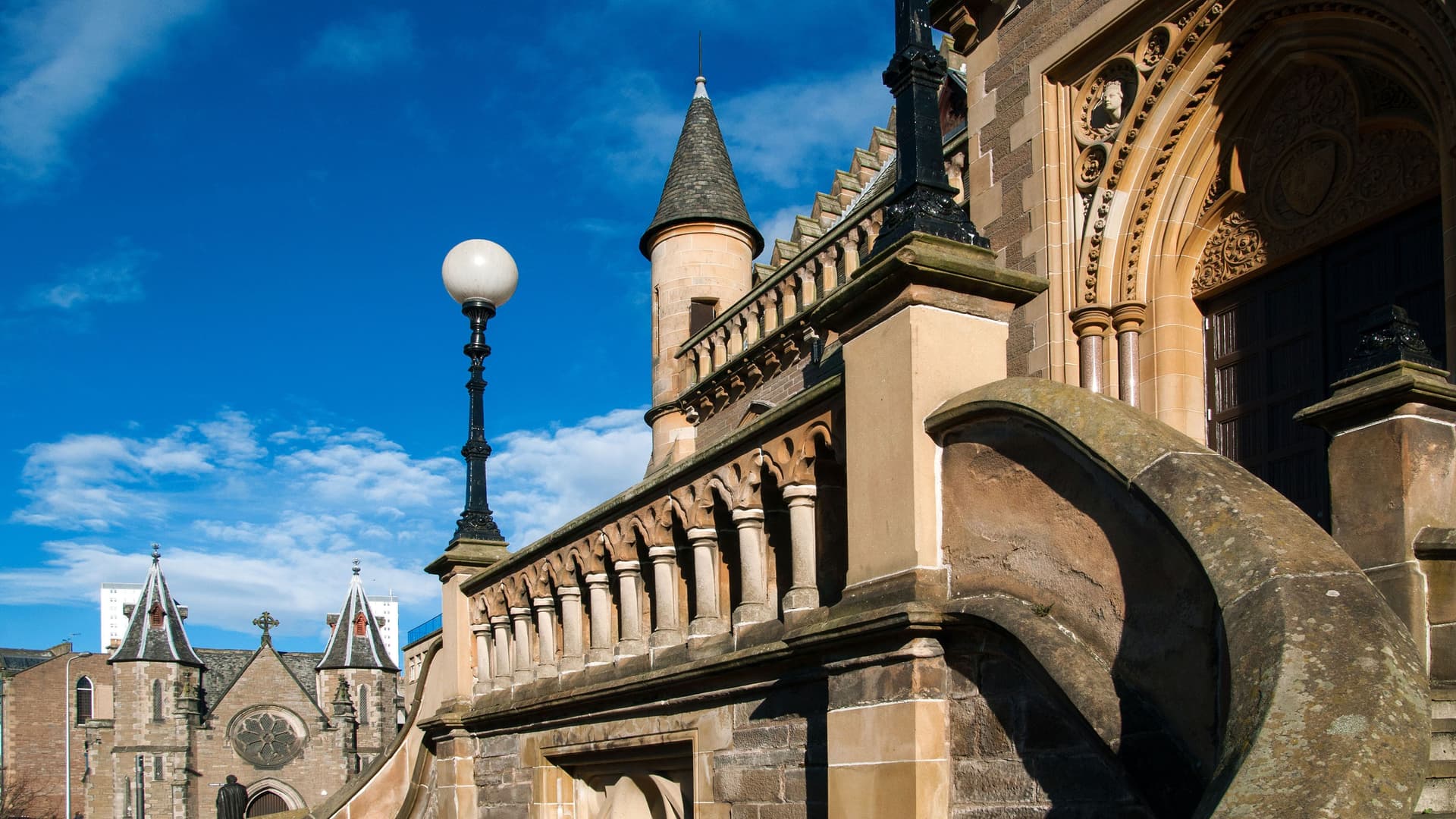 A historic stone building in Dundee, Scotland, with a large ornate archway, a turret, and a lamp post, set against a bright blue sky with white clouds.