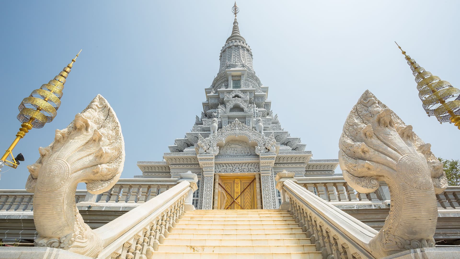 A stunning low-angle view of the ornate Udong mountain pagoda in Kampong Tralach, Cambodia, featuring a grand staircase flanked by guardian Naga snake statues leading up to the intricately decorated temple.