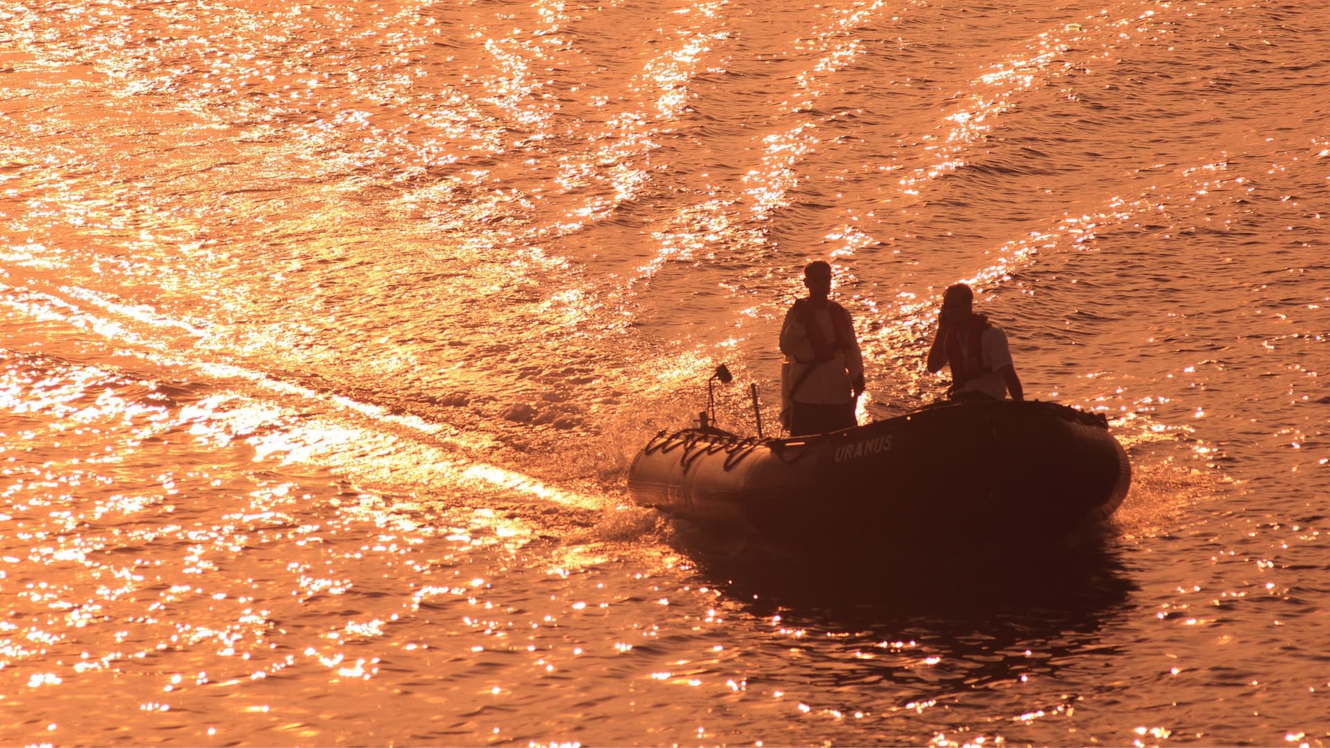 Two people ride a small rubber boat across the glittering, orange-colored water at sunset near Kenanga Village, Indonesia.