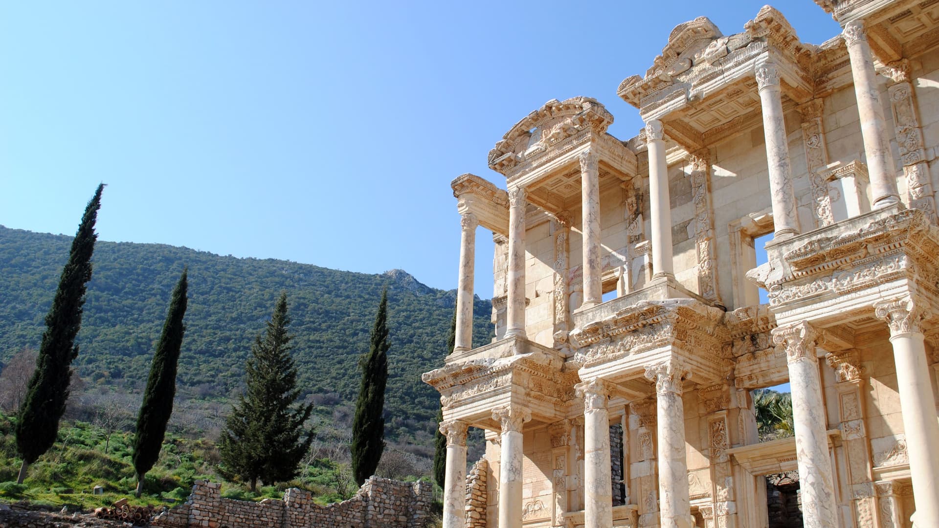 The iconic Library of Celsus in the ancient city of Ephesus, Turkey, with its magnificent, two-story facade and marble columns standing against a backdrop of cypress trees and green hills.
