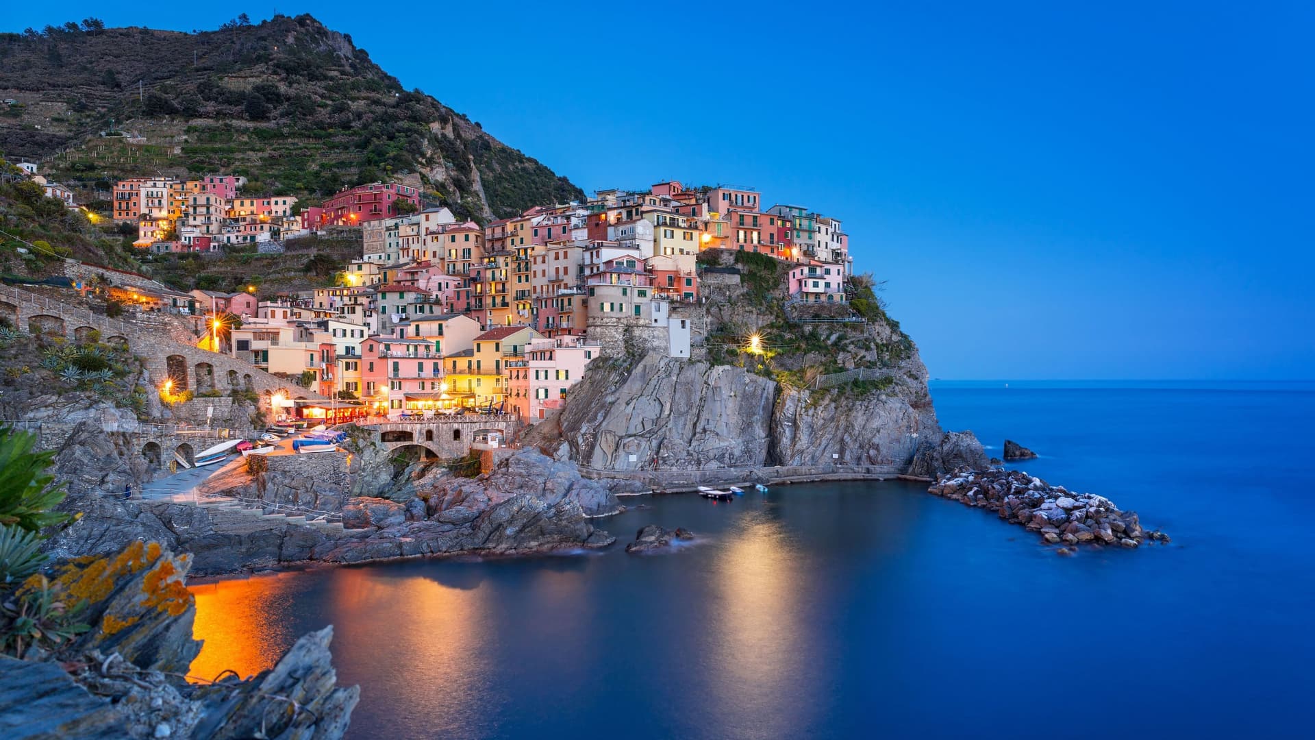 A breathtaking dusk view of the picturesque, colorful village of Manarola, a popular travel destination in Cinque Terre, Italy, with lights reflecting on the Ligurian Sea.