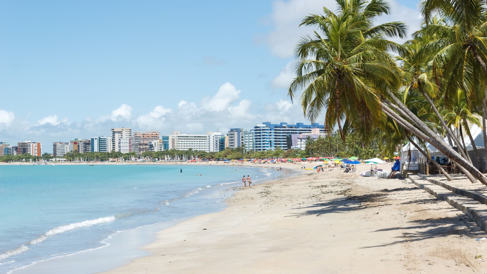A scenic shot of the beautiful Maceió beach in Brazil, with a calm turquoise sea, a stretch of golden sand, and tall palm trees lining the shore with the city skyline in the distance.