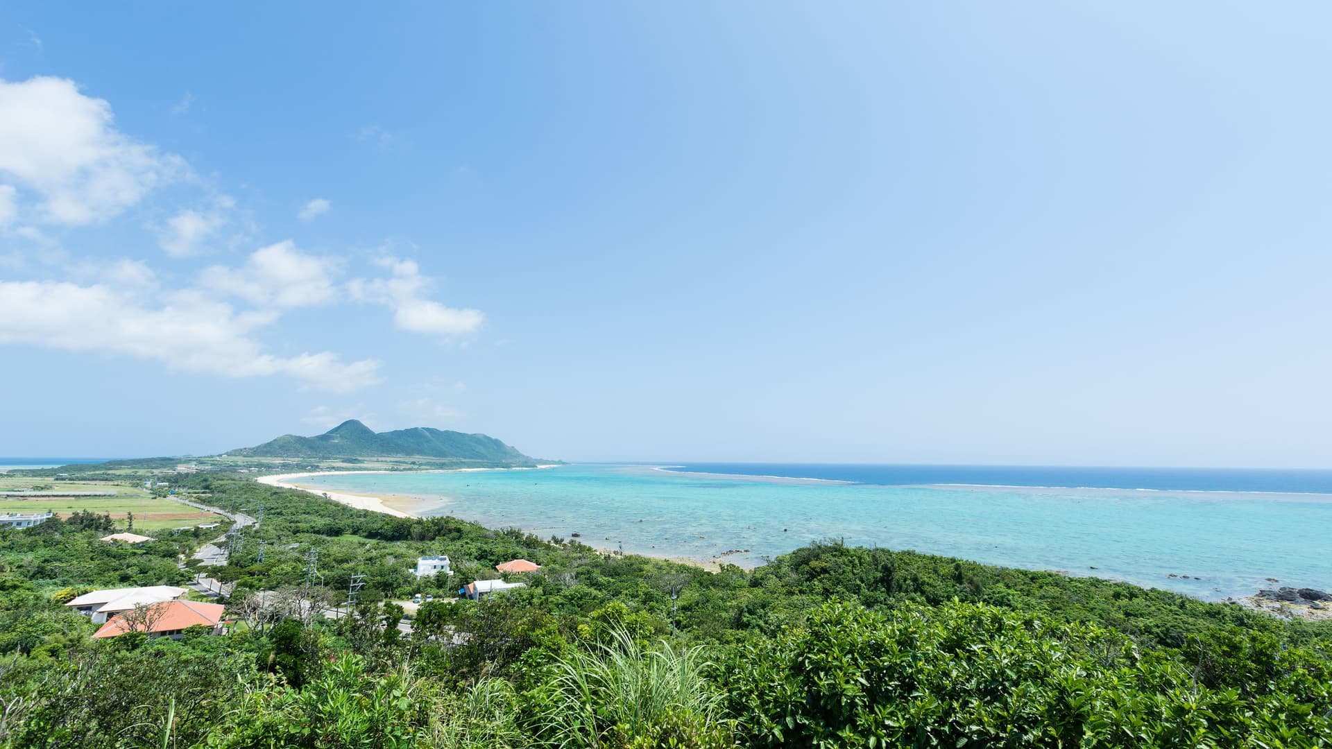 A stunning aerial shot of the coastline in Maizuru, Japan, with turquoise water, a sandy beach, and a lush green landscape under a clear blue sky.
