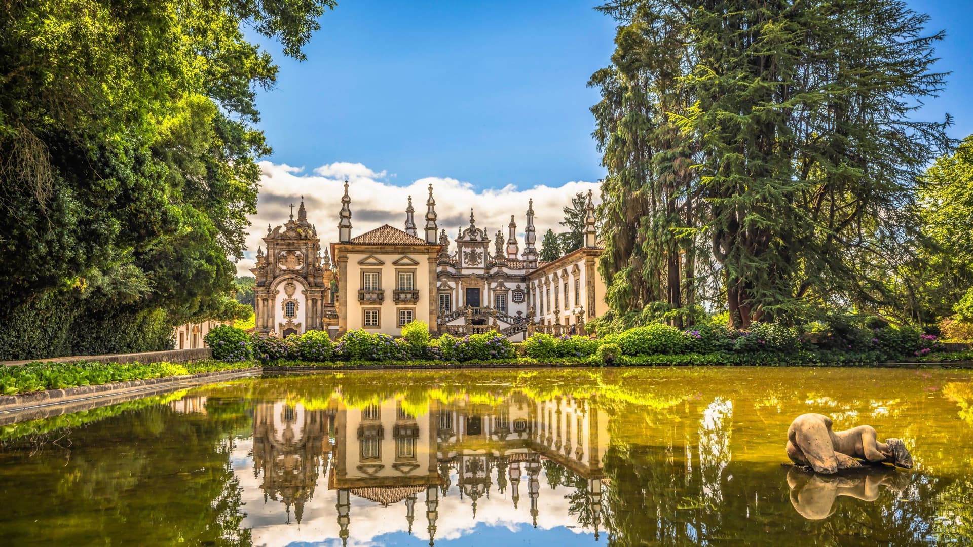 A picturesque view of the Baroque-style Casa de Mateus palace in Portugal, with its ornate facade and garden reflecting on a tranquil pond with a statue.