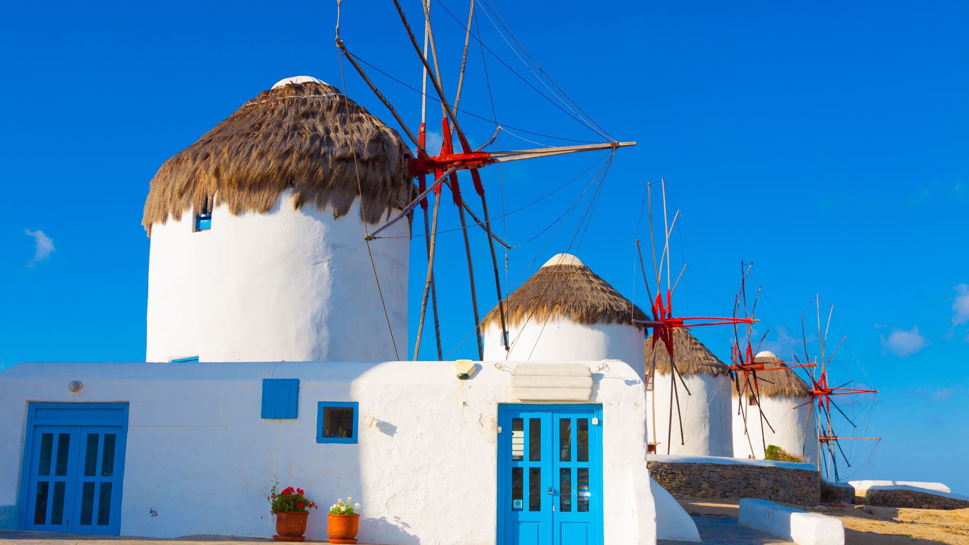 A beautiful daytime view of the iconic windmills of Mykonos, Greece, with their white-washed walls and thatched roofs against a vivid blue sky.