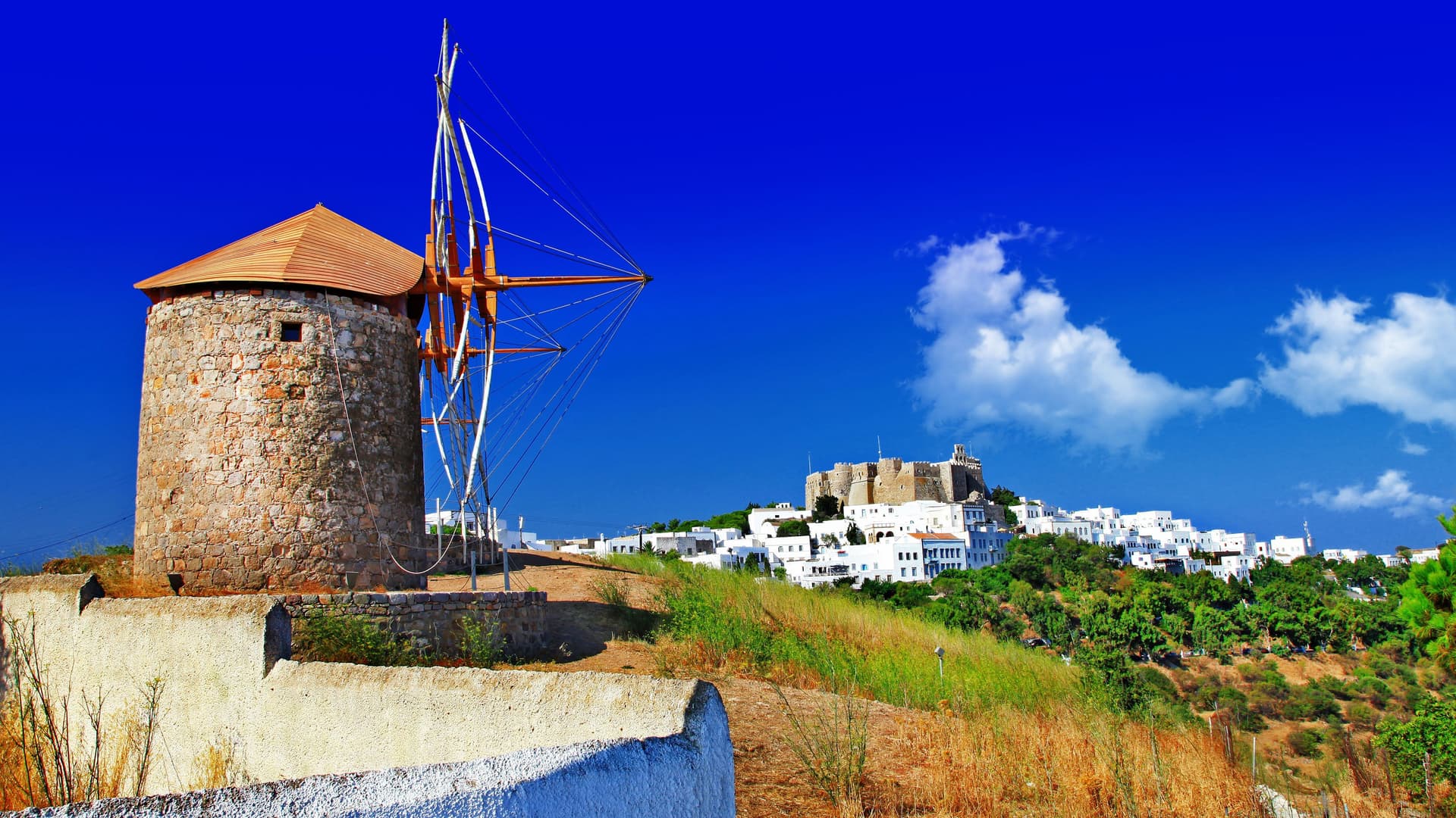 A traditional windmill with a pointed roof and wooden blades on a hill overlooking the white buildings of Patmos, Greece, with the historic Monastery of Saint John in the distance.