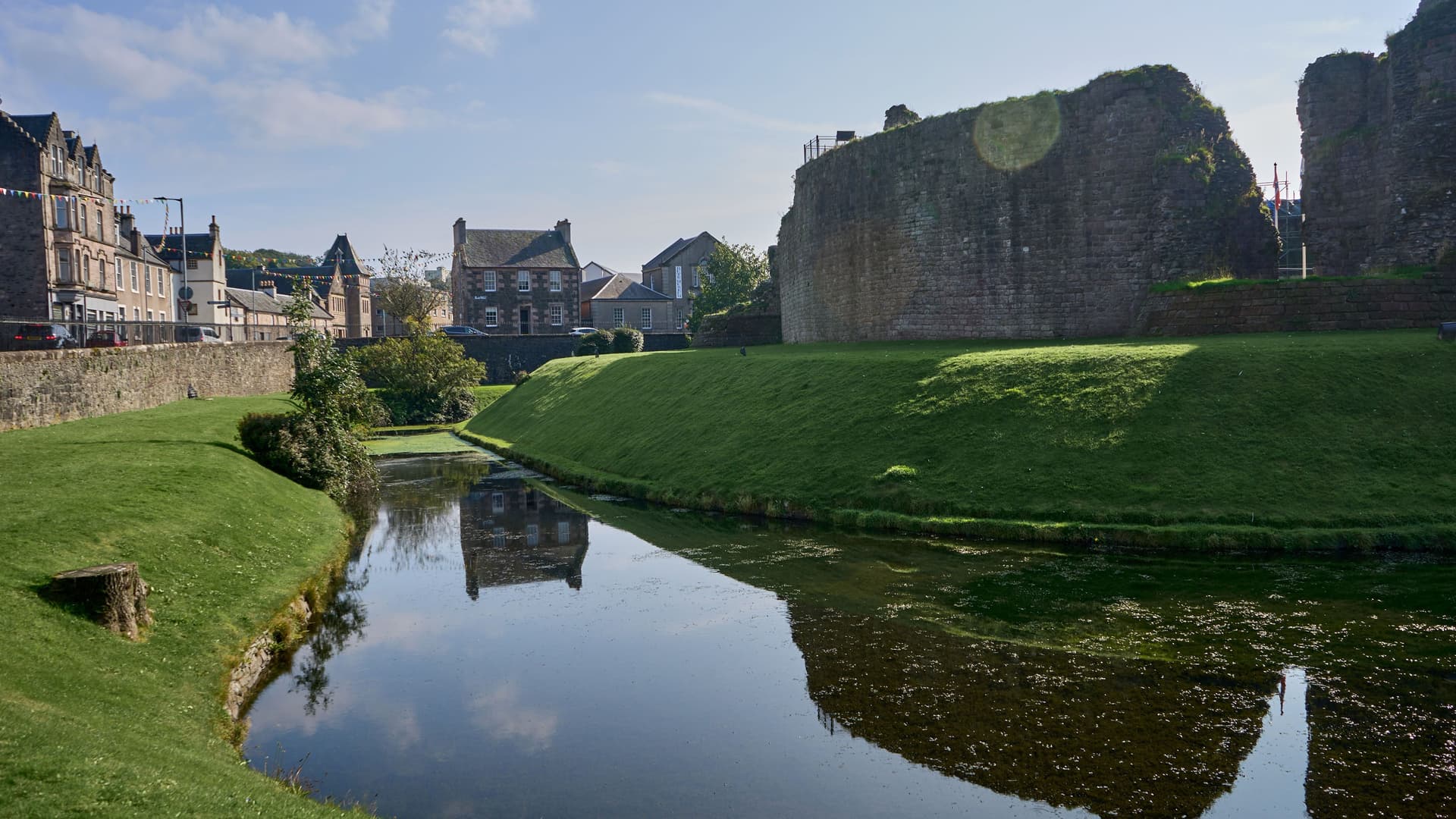 A picturesque view of the ruins of the medieval Rothesay Castle on the Isle of Bute, Scotland, with a tranquil moat and green lawn in front of historic stone houses.