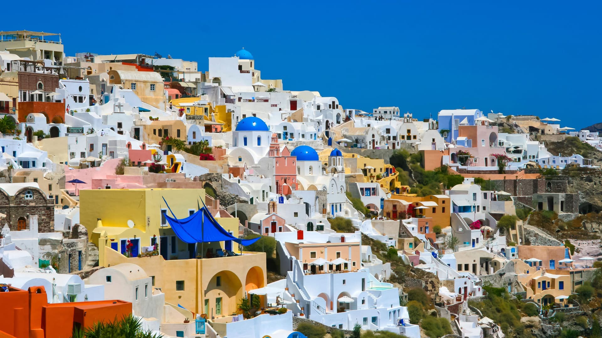 A stunning photograph of the iconic cliffside village of Oia in Santorini, Greece, with its whitewashed buildings and blue-domed churches cascading down the volcanic caldera, set against a deep blue sky.