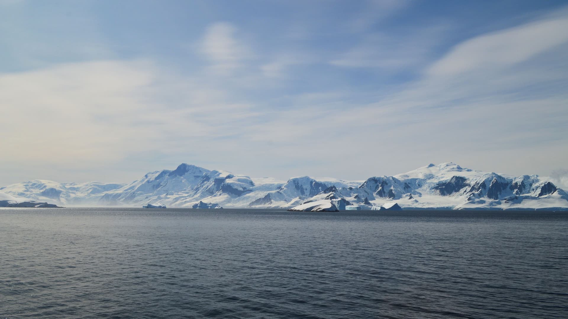 A panoramic view of the vast, icy landscape of Antarctica, with massive snow-covered mountains, glaciers, and icebergs floating in the deep blue waters of the Schollaert Channel.