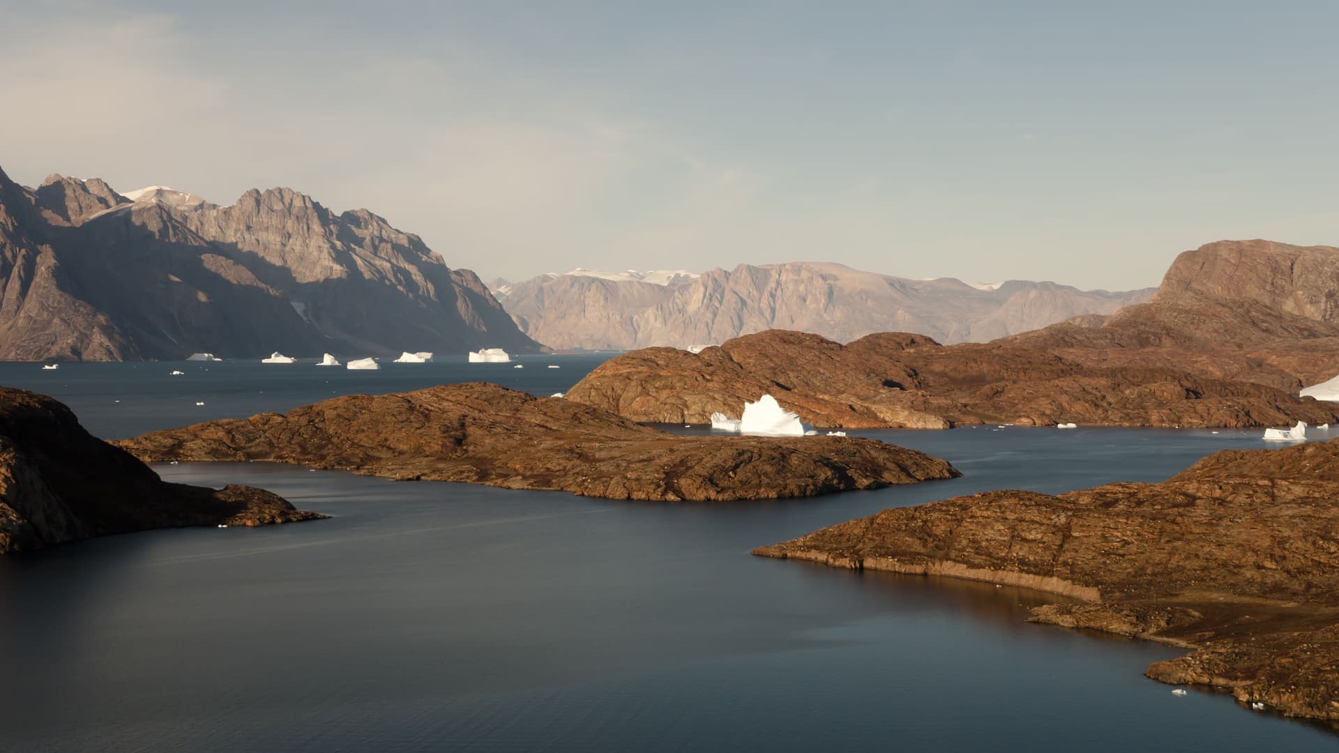 A breathtaking landscape shot of Scoresby Sund, Greenland, featuring floating icebergs and small brown islands surrounded by deep blue water and majestic mountains.