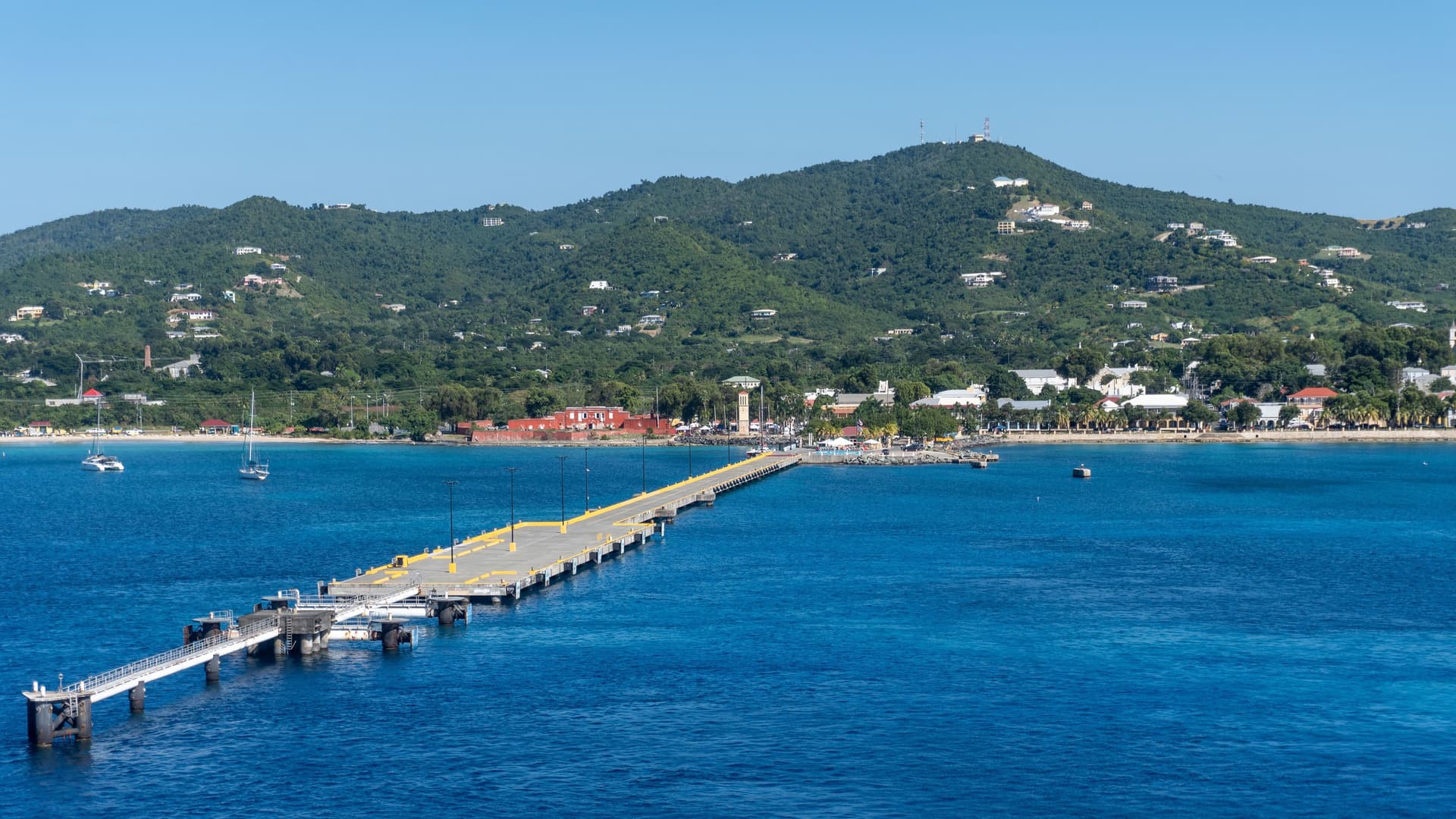 A panoramic view of a long, gray pier extending into the calm turquoise waters of St. Croix, US Virgin Islands, with the town and a lush green mountain in the background.