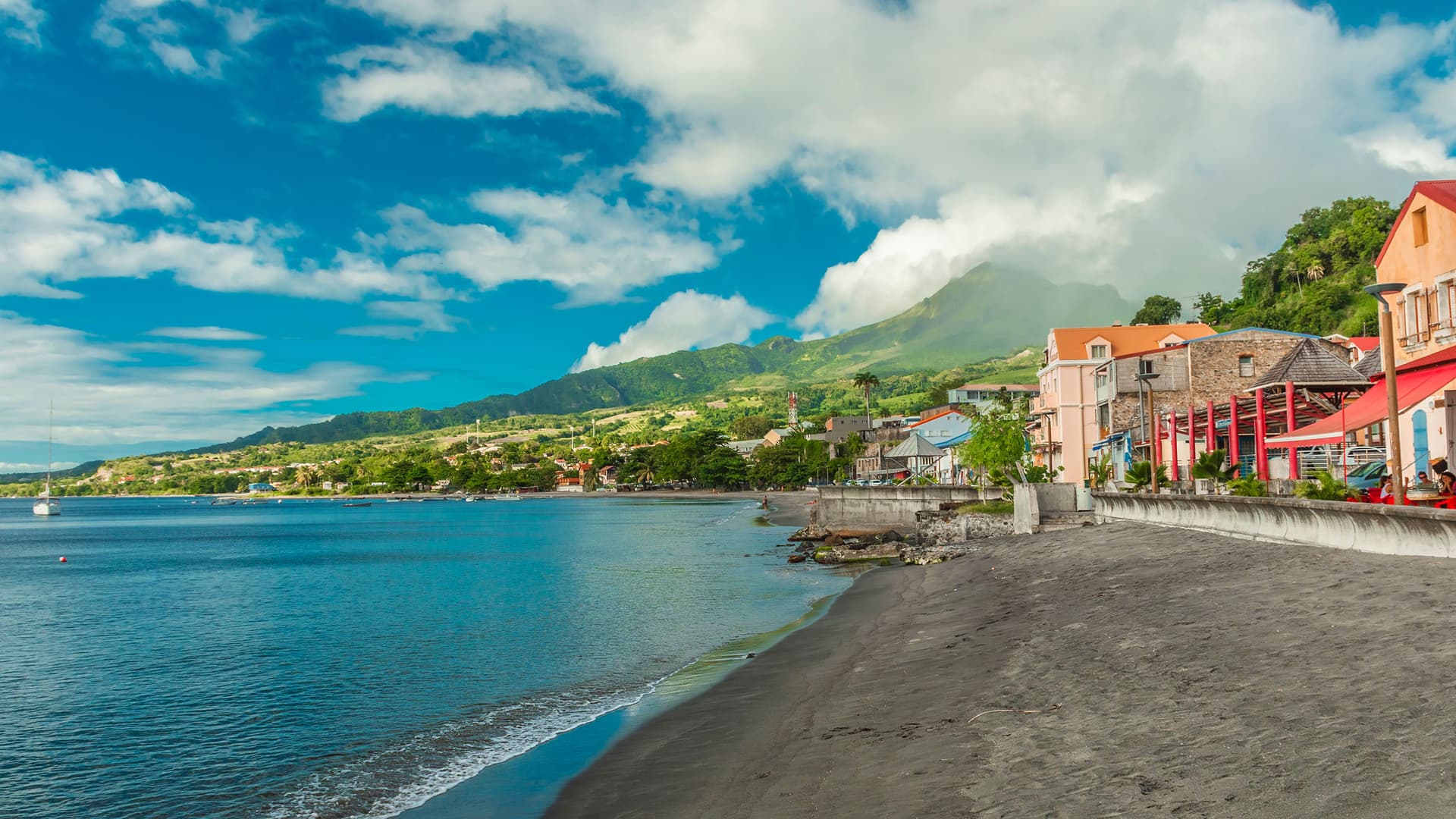 A view of the volcanic black sand beach and coastline of St. Pierre, Martinique, with colorful buildings and a lush, mountainous landscape under a beautiful sky.