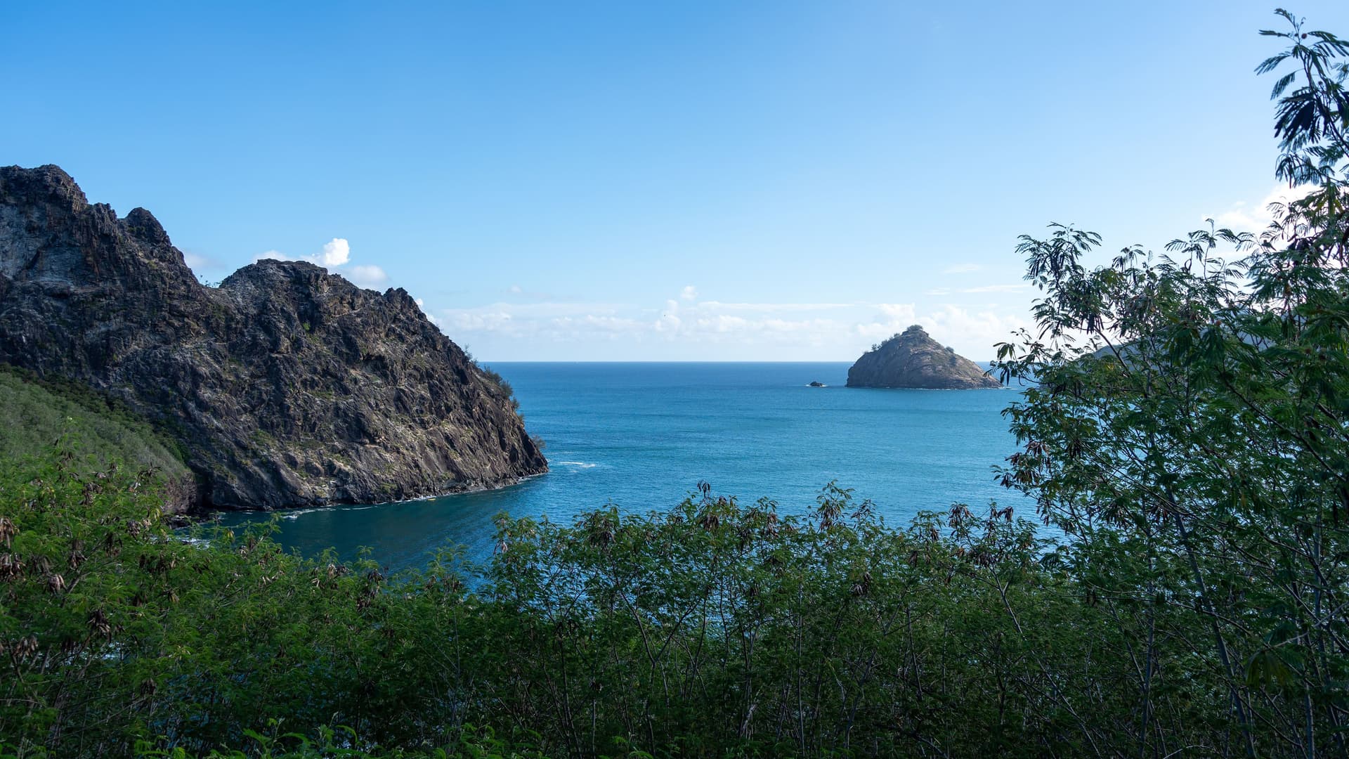 A stunning view of the deep blue waters of Taiohae Bay on Nuku Hiva in the Marquesas Islands, with rugged rock formations on the left and a small island in the distance. The foreground is filled with lush green foliage under a clear blue sky.