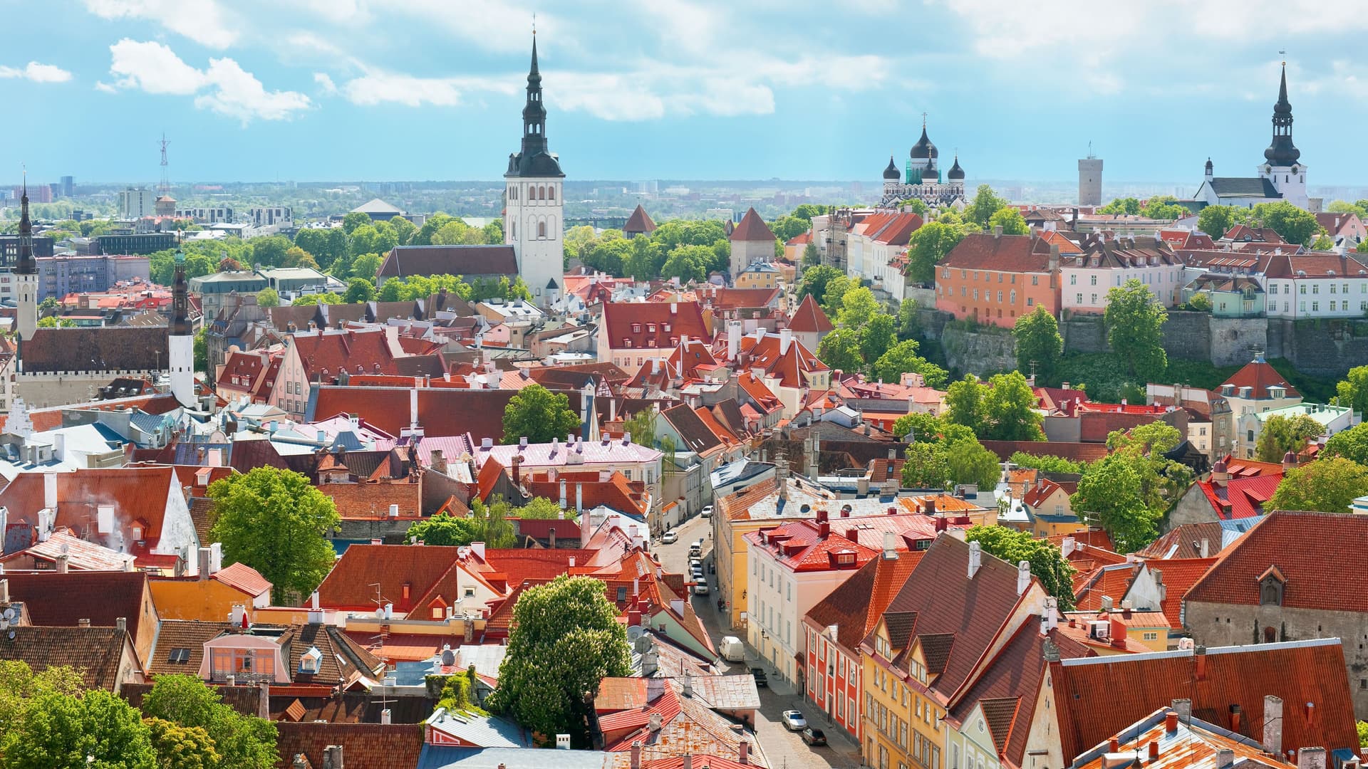 An expansive panoramic view of the medieval Old Town of Tallinn, Estonia, with its iconic red rooftops and historic church spires against a bright, cloudy sky.