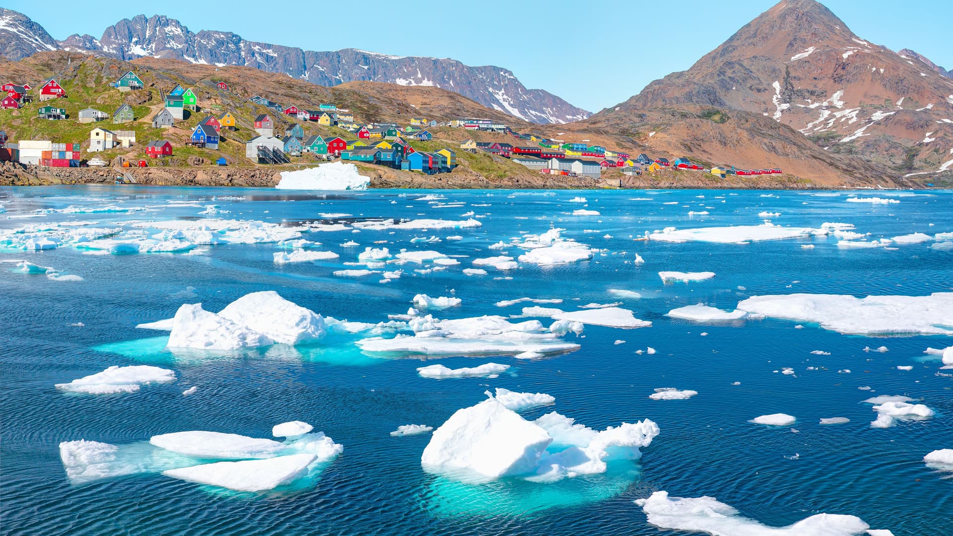"A colorful collection of houses in Tasiilaq, Greenland, built on a rocky shoreline and a hillside overlooking the deep blue water of the Arctic Ocean, dotted with white icebergs and floating ice.  "