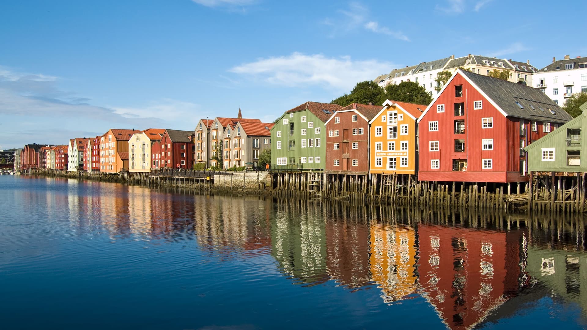 A scenic view of historic, colorful wooden warehouses built on stilts along the Nidelva River in Trondheim, Norway, with their reflections visible in the calm water.