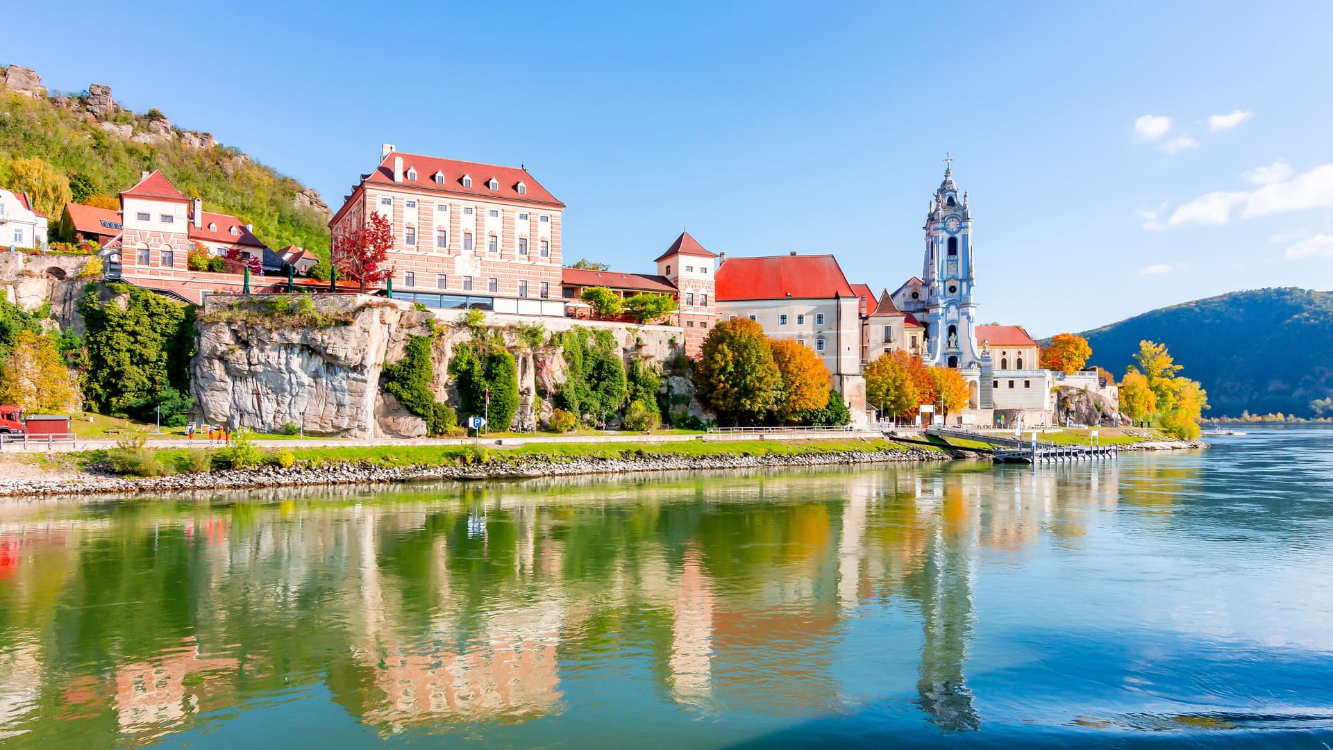 A sunny view of the historic town of Dürnstein in the Wachau Valley of Austria, with its famous baroque church and red-roofed buildings reflecting in the calm waters of the Danube River.